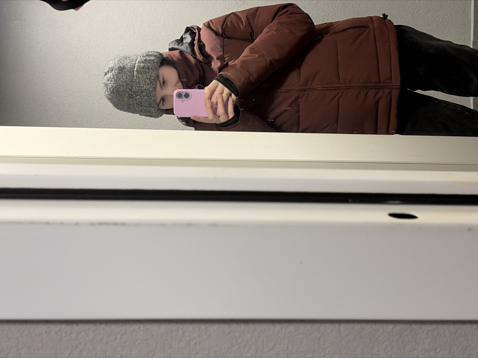 The scene depicts an individual, appearing female, captured in a mirror reflection within an indoor environment in the city of Ковалёво, Russia. The person is shown from mid-thigh up, wearing a grey knit beanie and a high-collared, rust-red or maroon puffer jacket over dark trousers. She holds a light pink smartphone, equipped with dual rear cameras, in both hands at chest level, positioning it to capture her reflection in the mirror. A ring is visible on the ring finger of her left hand. The immediate background behind the reflected person is a plain, light grey wall. The left side of the image prominently features a white door frame or trim, which partially obstructs the view of the reflection. This frame includes a vertical dark strip or seal and a small, dark oval-shaped opening. The wall surface adjacent to the white frame on the far left exhibits a textured, muted grey finish.
