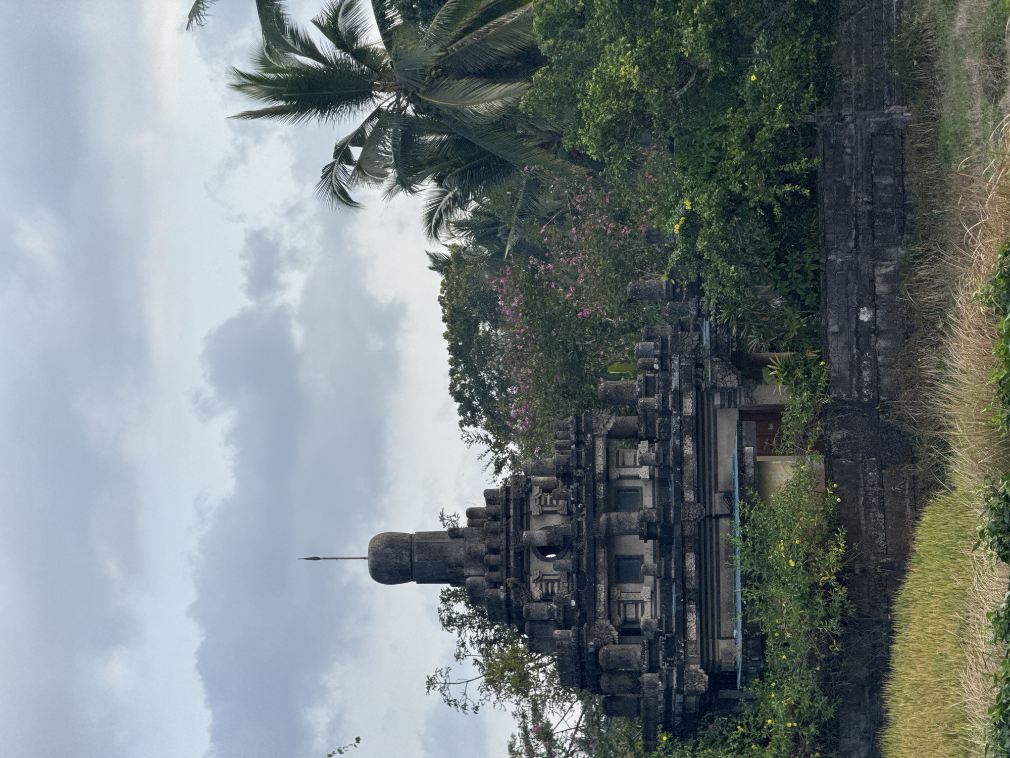 A weathered, stone temple structure stands amidst lush greenery, characteristic of Tegallalang, Indonesia. The temple features a layered, tiered design with intricate carvings and decorative elements, topped by a finial. It is partially obscured by dense foliage, including trees with vibrant pink flowers and towering palm trees. In the foreground, dry grasses and a field of crops suggest an agricultural setting. The sky is overcast with soft, diffused light. There are no people visible in the image. The scene captures a tranquil, possibly ancient, architectural site integrated into a tropical landscape.