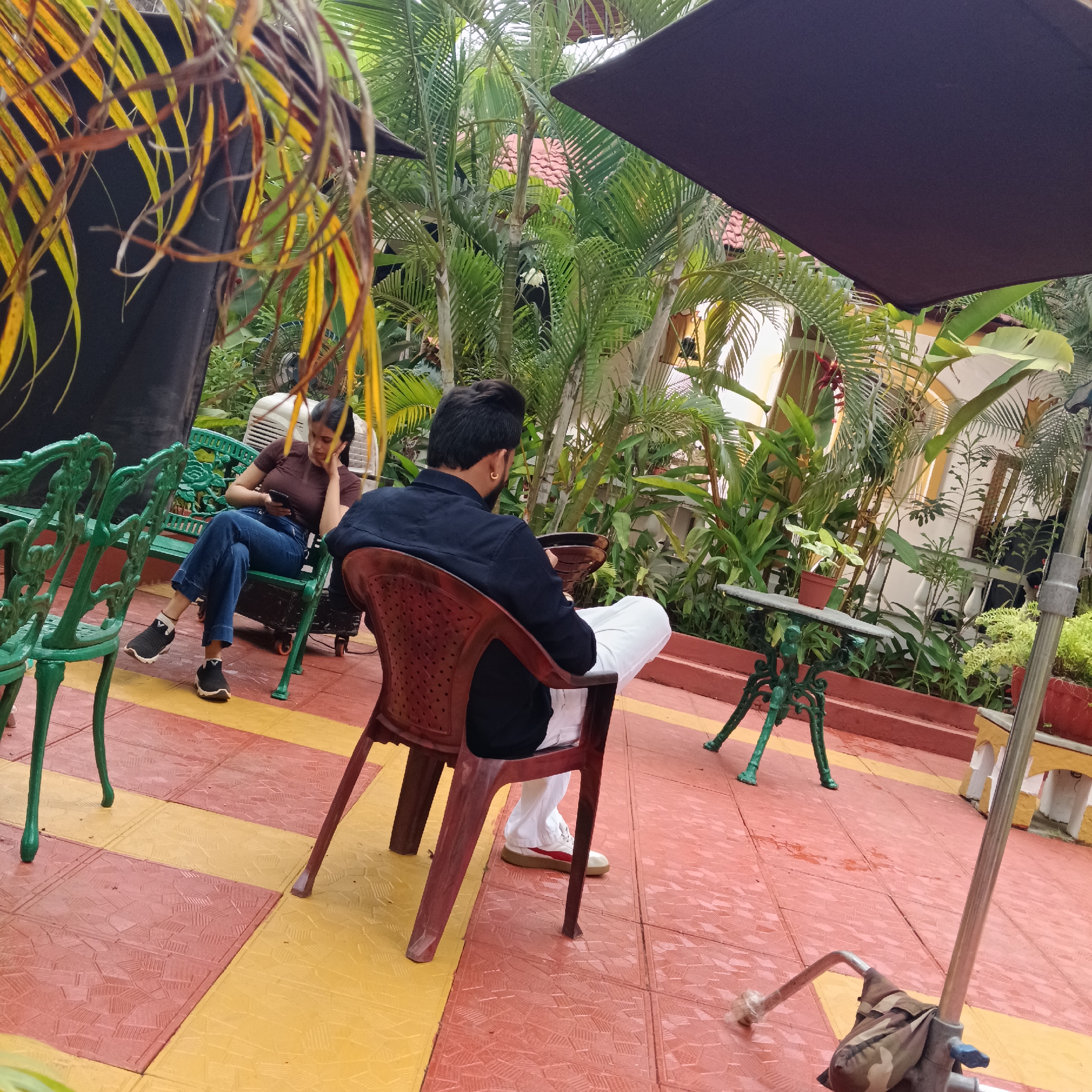 The image captures a scene in what appears to be a tropical courtyard or garden in Anjuna, India. A woman sits on a green wrought-iron bench, looking at her mobile phone. She is wearing a brown top, blue jeans, and black sneakers. A man sits in a brown plastic chair facing away from the camera, wearing a black shirt and white trousers with white and red sneakers. The ground is tiled in a red and yellow checkered pattern. Tropical plants and trees, including palms, fill the background. A black umbrella-like object is positioned overhead, possibly for shade or as part of a filming setup. Green wrought-iron tables and chairs are scattered around. There is also a metal stand with a camo bag on the lower right. The environment appears to be a relaxed outdoor setting, perhaps a cafe or garden within a hotel.
