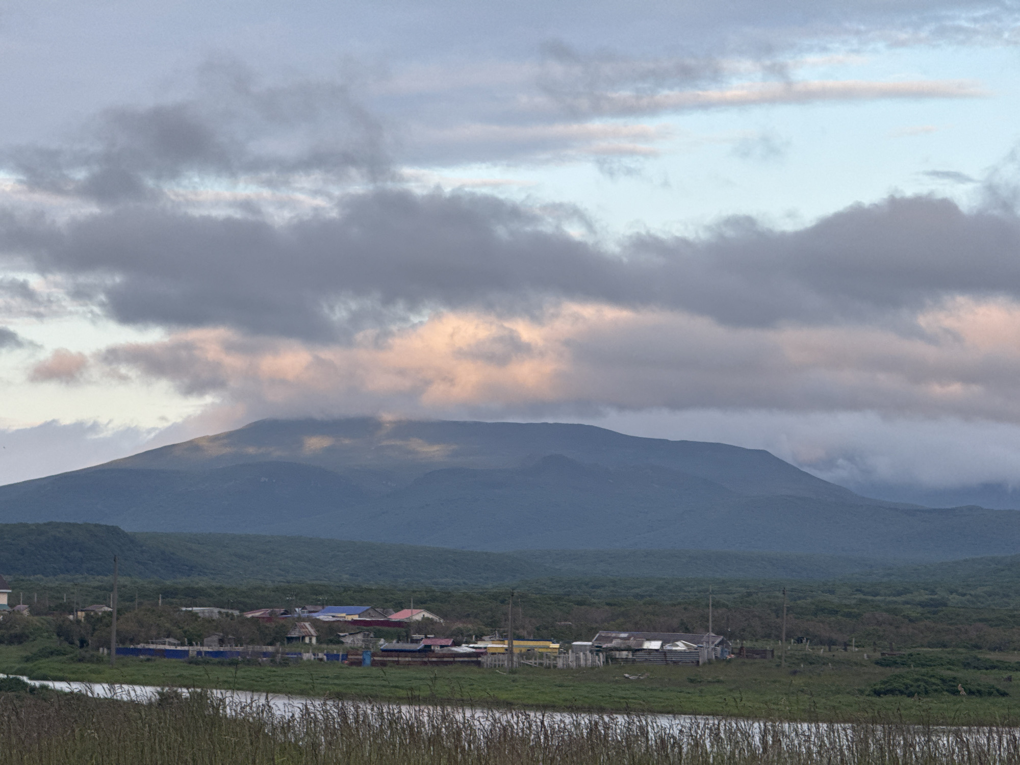 An overcast sky with patches of pinkish light above a muted, hazy mountain range. In the mid-ground, a cluster of small, weathered buildings with multi-colored roofs forms a village. The village is situated on a grassy plain, with a body of water visible in the foreground, bordered by tall reeds. The scene is tranquil and appears to depict a rural coastal area. The location is the city of Kuril'sk, Russia.