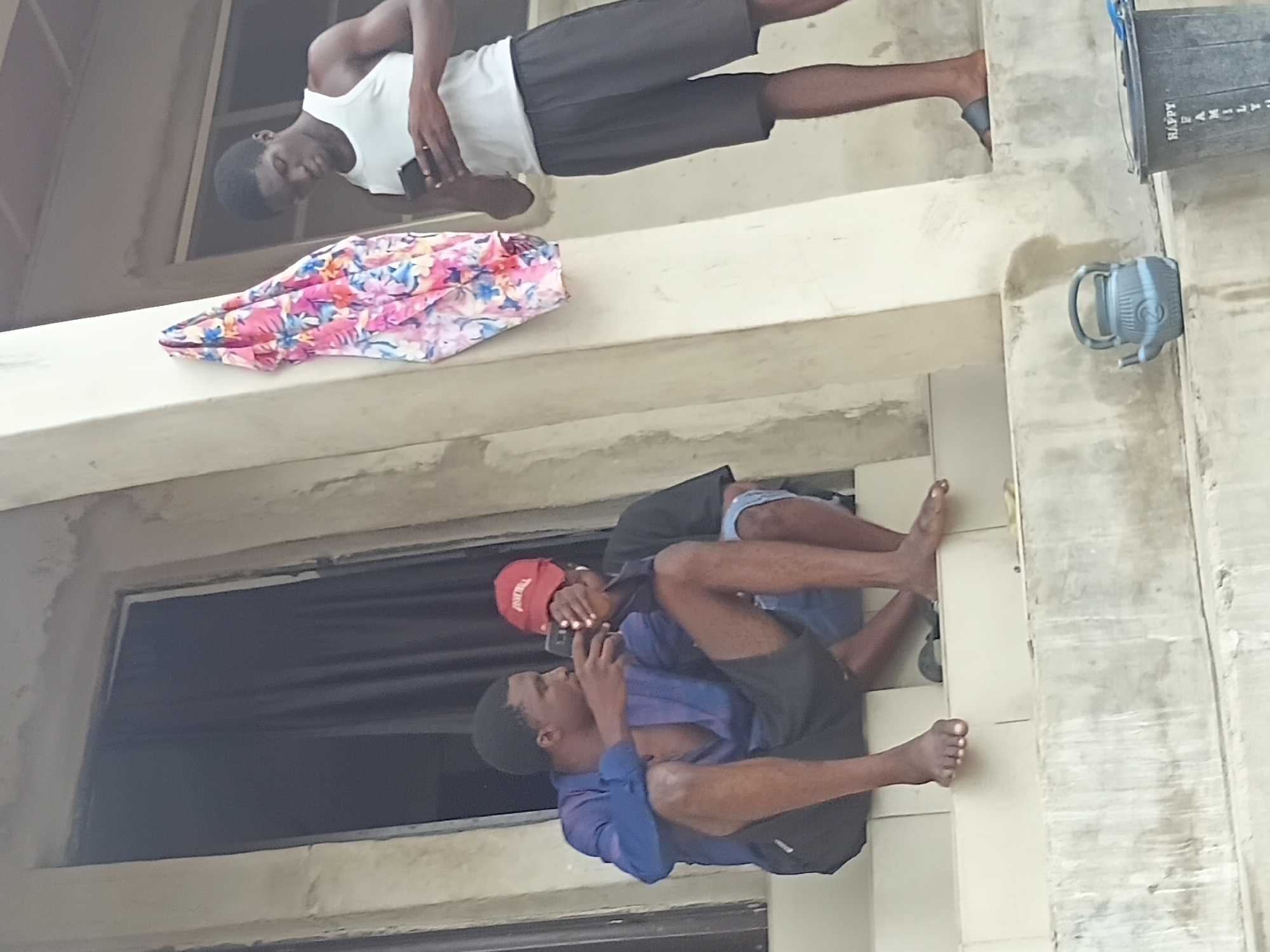 A medium shot shows three individuals on a building exterior in Unknown, Unknown. The building has a multi-level concrete structure with window openings. A male with dark hair, wearing a white tank top and dark shorts, stands on an upper level. A floral-patterned cloth rests on the concrete near him. Below, two other males stand on a lower level, one wearing a red baseball cap and a blue shirt, while the other wears a black shirt. Both wear shorts and are barefoot. One of them appears to be holding a phone to the ear of the other. A gray-blue bucket is seen attached to the building's right-side edge. Another larger container, partially visible, sits atop the structure.