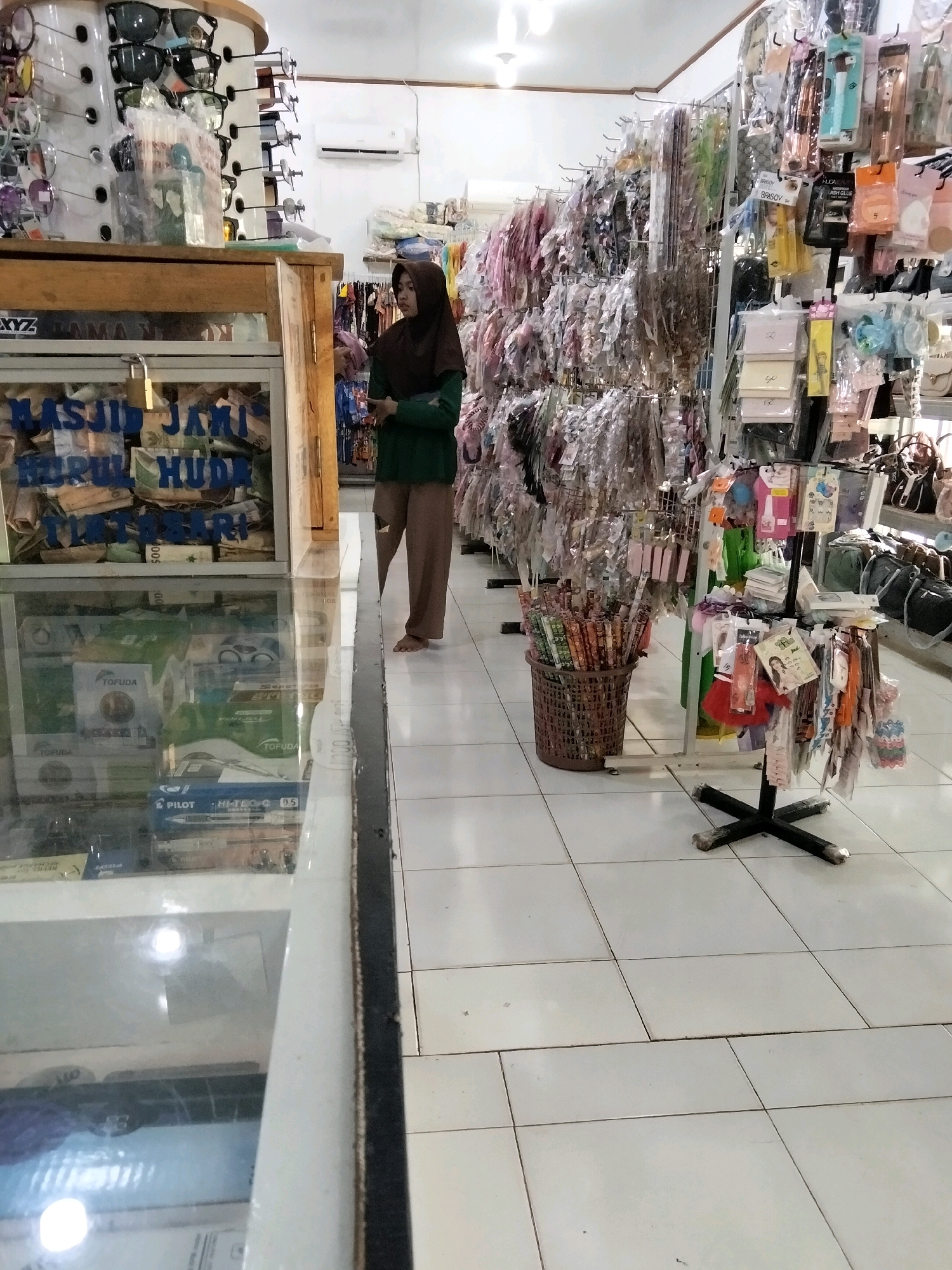 The image depicts the interior of a retail store, possibly a small shop or kiosk, located in Tirto Sari, Indonesia. The store appears to sell a variety of small goods and accessories, with shelves and racks filled with packaged items such as cosmetics, stationery, and eyewear.
In the foreground, a glass display counter shows various items, including what appear to be pens and stationery products. Behind the counter, a young woman wearing a dark brown hijab and a green long-sleeved shirt stands and observes. Her lower body is visible, clad in brown trousers, and she is barefoot.
The store's walls are lined with shelves displaying an abundance of merchandise. A rack of sunglasses is prominently displayed on the left side of the image. To the right, an extensive display of packaged accessories, likely hair accessories, makeup, and small trinkets, is arranged on metal racks. A basket filled with rolled items, possibly umbrellas or posters, sits on the floor to the left of the woman. The floor is tiled in a light, off-white color. The overall impression is of a busy, well-stocked retail environment.