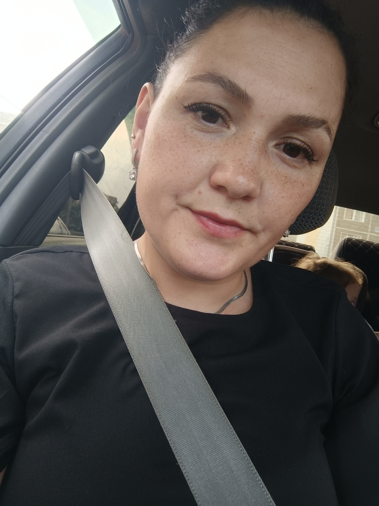A close-up, selfie-style shot shows a woman with dark hair pulled back, wearing a black shirt and a seatbelt. She has fair skin with freckles and is wearing a dangling earring in her left ear. Her mouth is slightly open, and she appears to be looking at the camera. The background suggests she is in a car, with a gray seatbelt diagonally across the frame and part of a car's interior visible. Behind her, another person's head with blonde hair is partially visible. The image appears to have been taken during the daytime, with bright light coming from the left. While the exact location within Chaikovsky, Russia is not discernible from the image alone, the context of being in a car implies a road or travel setting within the city.