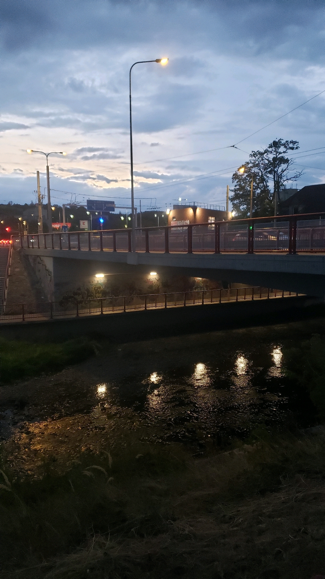 The image depicts a bridge spanning a body of water at dusk in the city of Prešov, Slovakia. The bridge, with a red railing, is illuminated by streetlights. Two levels of the bridge are visible, with the upper level carrying traffic, indicated by the red and yellow lights of vehicles on the left side of the frame. The lower level of the bridge features a pedestrian walkway with lights. Reflections of the bridge lights are visible on the surface of the water below. The sky is cloudy and displays the transition from daylight to twilight. Vegetation, including grass and reeds, lines the banks of the waterway. The overall scene captures a quiet urban evening with elements of infrastructure and nature.