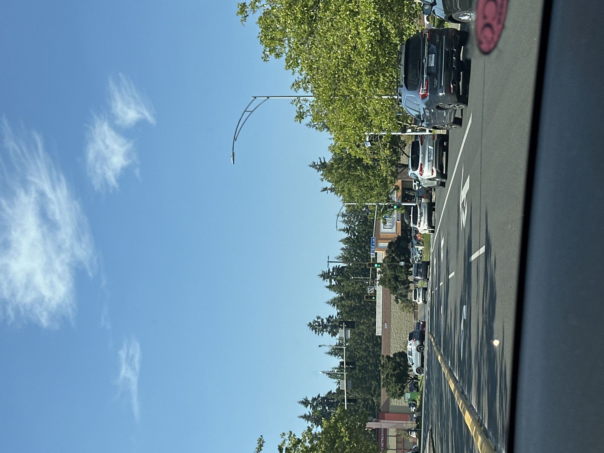 The image shows a daytime view of a multi-lane road in Lynnwood, Washington, United States of America. The road is lined with trees and buildings, including a US Metro Bank. Cars are visible driving in the same direction as the camera, with traffic lights and street signs indicating an intersection. The sky is clear blue with scattered white clouds.
