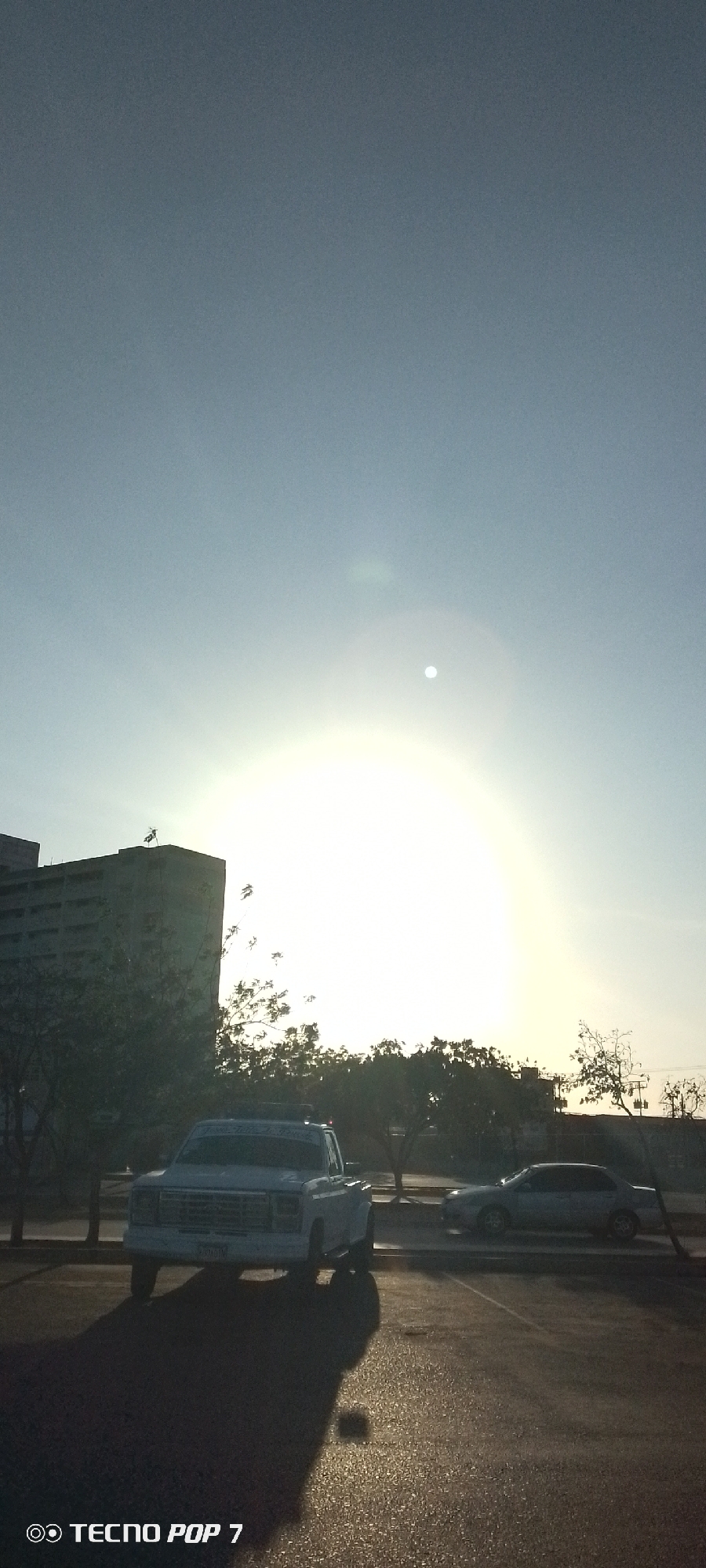 The image shows a parking lot in Juan Griego, Venezuela at dawn. A light-colored pickup truck is parked in the foreground, casting a long shadow. A building is partially visible to the left, and trees line the background. A silver sedan is parked behind the truck. The sun is bright, obscuring details in the middle distance. The camera model is TECNO POP 7.