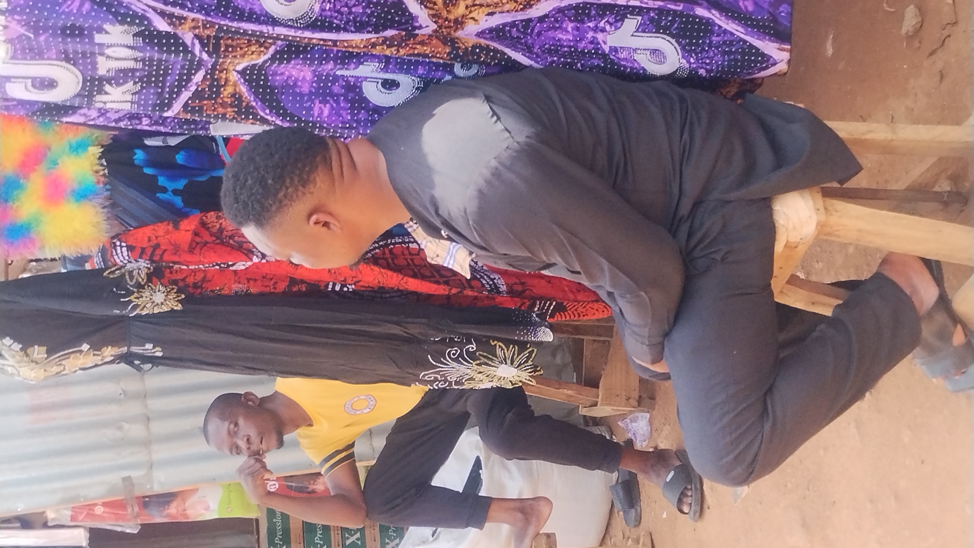 The scene depicts two men in an outdoor setting. One man, dressed in a black shirt and trousers, is seated on a simple wooden stool. The second man, wearing a yellow shirt and black trousers, is standing nearby, appearing to pick his teeth. Behind the men are various colorful fabrics and textiles, suggesting a market or shop environment. In the foreground, there is a sign displaying the word "X-Presslon." The location is likely in Jos, Nigeria, based on the cultural context of the scene. The ground appears to be dirt.