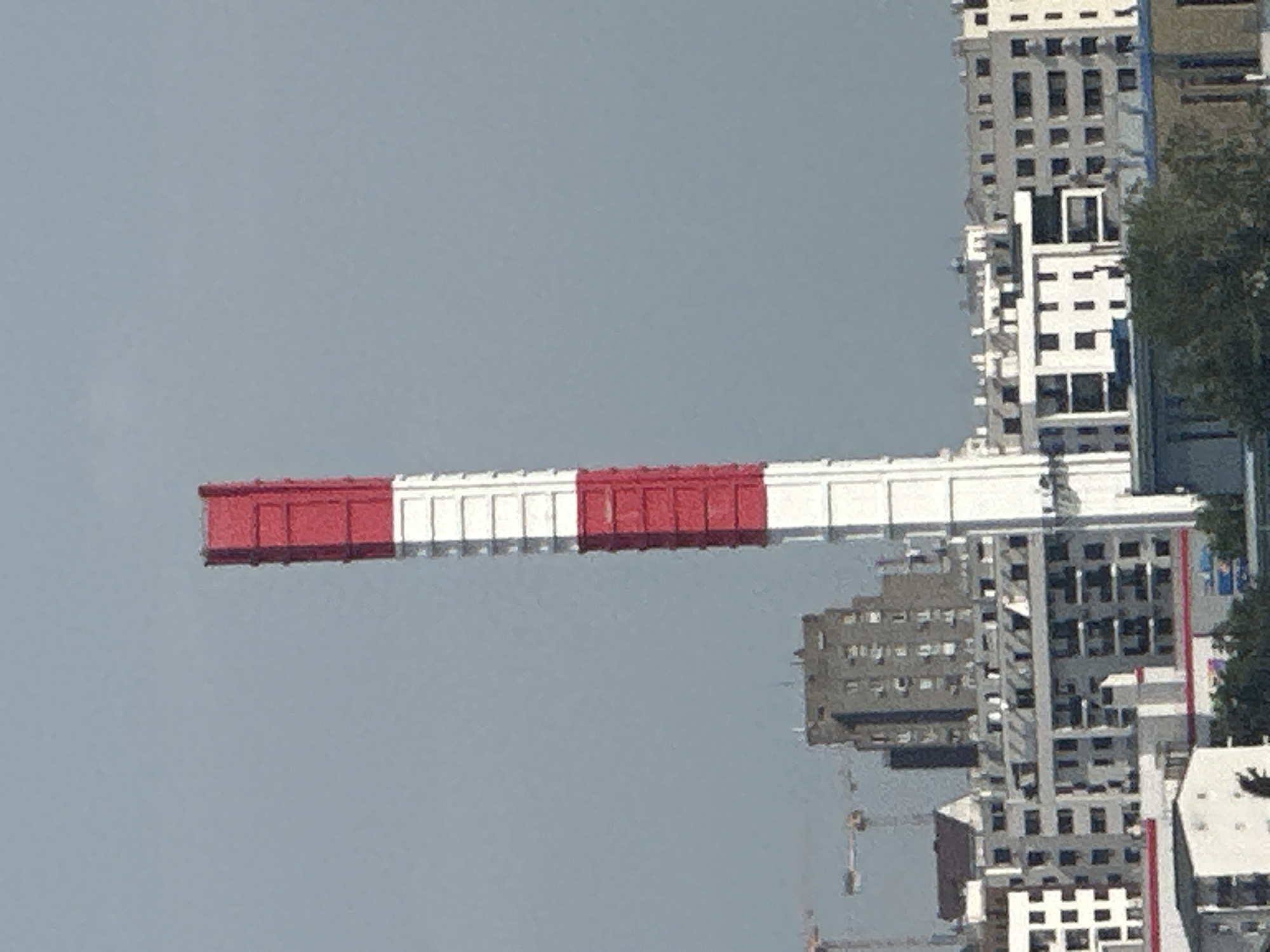 The image depicts a tall, striped chimney, red and white, standing against a hazy sky. The chimney is the most prominent object, leaning slightly to the left. Below and behind the chimney, a dense urban landscape is visible, characterized by a variety of buildings. These include mid-rise apartment buildings with numerous windows, some with balconies, and a few taller, more monolithic structures. Construction cranes are also present in the background, suggesting ongoing development. The overall scene is one of an industrial or mixed-use area within a city. Based on the characteristic architecture and the presence of similar chimneys and urban planning, this image is likely located in Belgrade, Serbia, possibly in the industrial zone of Zemun or near the former industrial complex of Dunav.