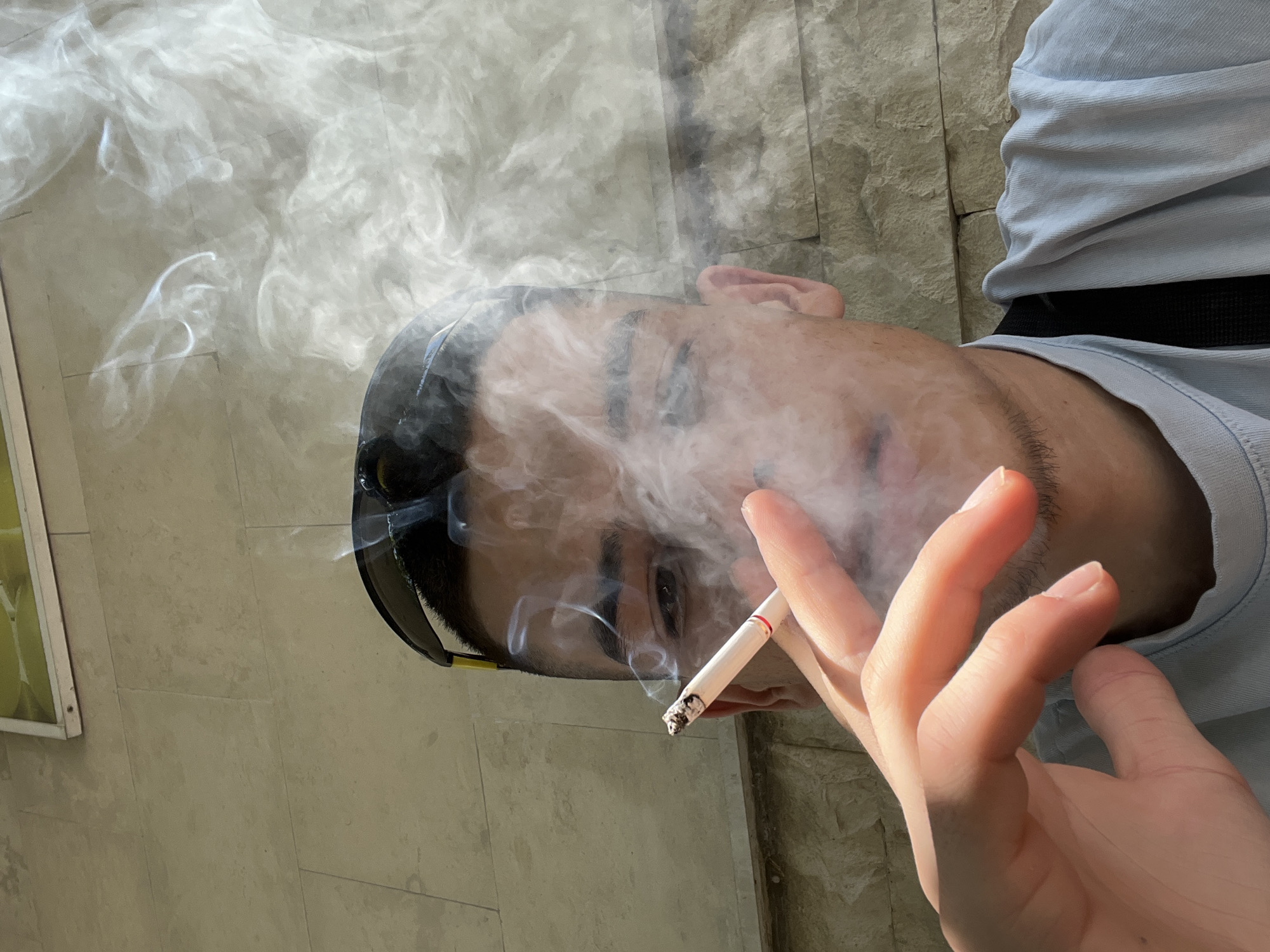 In the city of Karlovo, Bulgaria, a close-up, eye-level, medium shot captures a young man smoking a cigarette. He is positioned in the foreground, with his face turned slightly towards the camera, partially obscured by wisps of smoke. His left hand is raised, holding the cigarette between his index and middle fingers. The man is wearing sunglasses perched on his head and a light blue t-shirt. The background consists of a textured stone wall. The lighting suggests an outdoor setting with sunlight casting shadows.