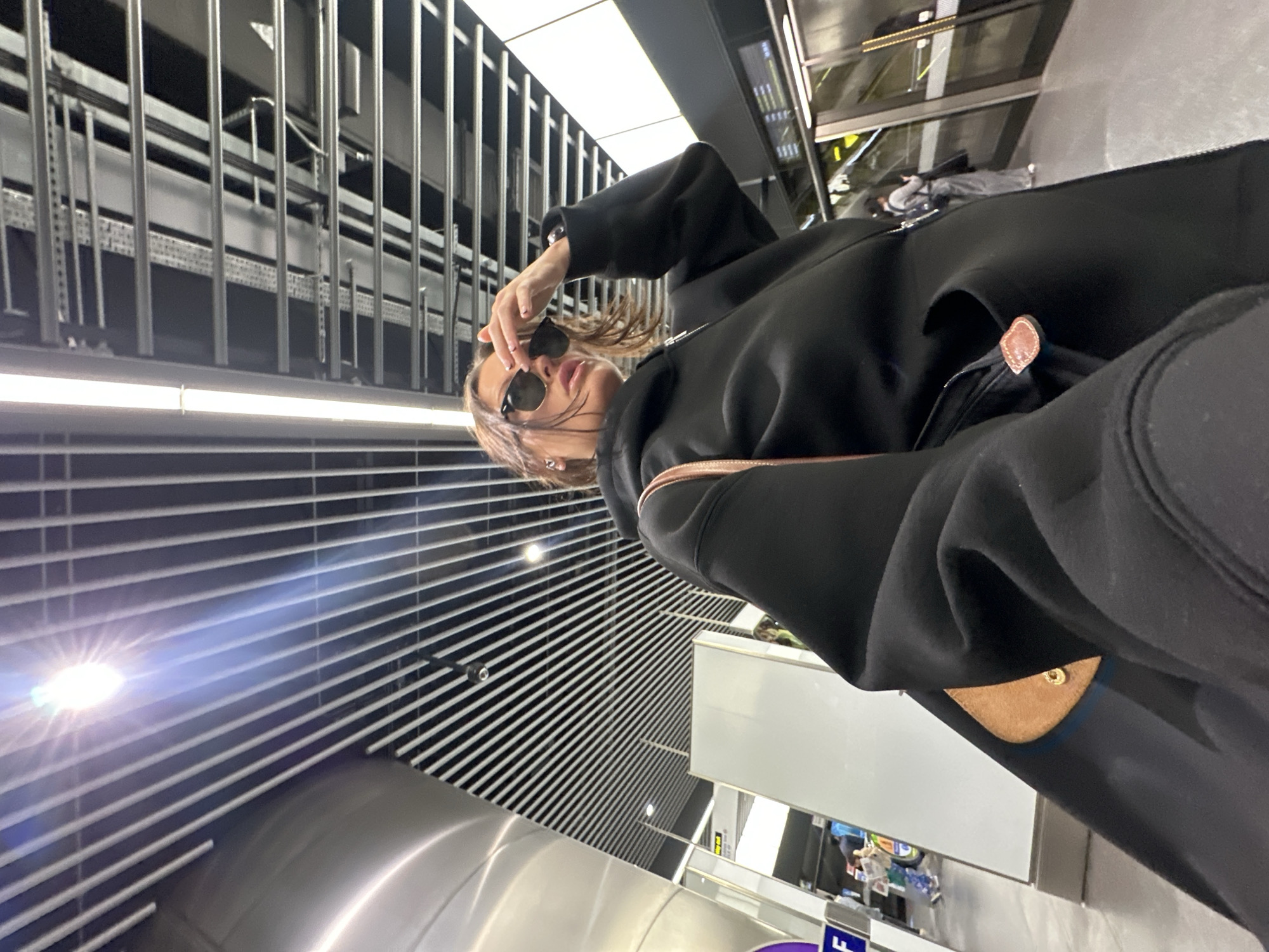 A medium shot captures a woman in a black hoodie and sunglasses, holding her sunglasses near her face. She appears to be in an underground station, likely in London, United Kingdom. Above her is a structure of linear metal bars. The background consists of the station platform, illuminated signage, and people waiting.