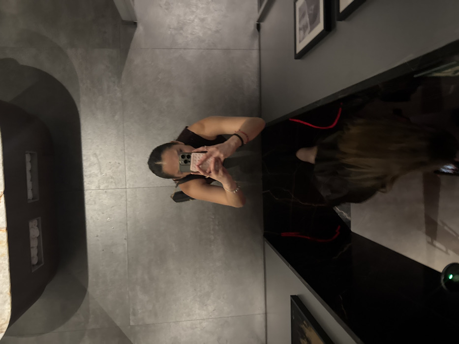 A woman stands on gray tiled flooring, taking a photo of herself in a bathroom mirror. She wears a dark top and holds a pink cell phone. The countertop is black with red trim. Framed artwork hangs on the wall behind the counter. A sink basin is partially visible on the left side of the image. A small electronic device is visible on the right side of the counter.