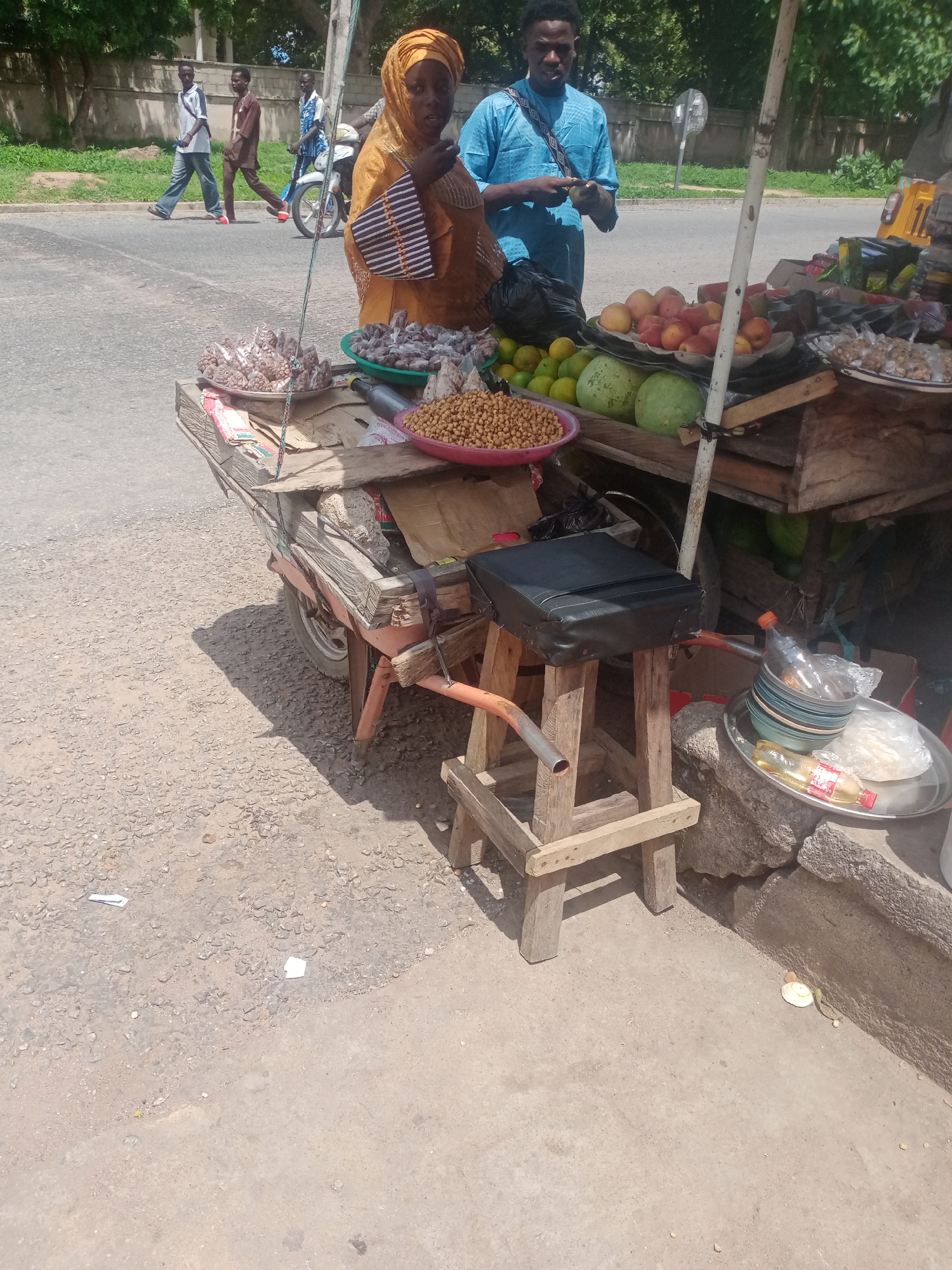 A street vendor's stall is set up on a roadside in Jalingo, Nigeria. The stall is a wooden cart laden with various goods, including fruits such as oranges and apples, and bags of dried produce. A woman in a bright orange headscarf and dress stands behind the stall, attending to customers. A man in a blue shirt is also present, interacting with the vendor. In the background, people are walking and a motorcycle is parked nearby. The scene depicts a typical market setting in a Nigerian city, with a focus on the vibrant colors of the produce and the clothing of the people. The stall is positioned on a dusty, unpaved surface, typical of many urban areas in Nigeria.