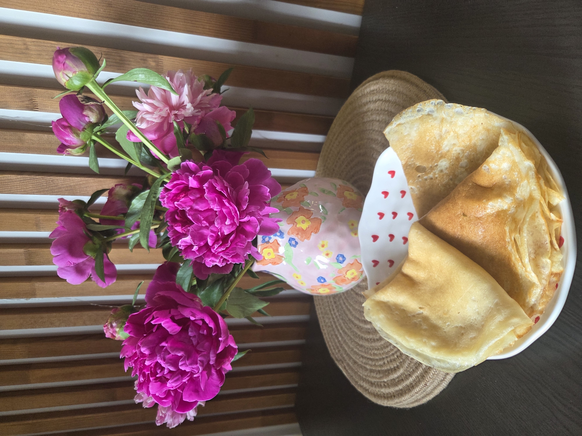 A still life image features a bouquet of vibrant pink peonies in a decorative vase, positioned on a woven placemat. The vase, with a light pink base and floral accents, sits on a dark brown surface. In front of the vase, a white bowl filled with folded crepes is visible. The background consists of vertical wooden slats. The image appears to be taken indoors, likely in a domestic setting, and contains no identifiable landmarks or clues to a specific location in Moscow, Russia.