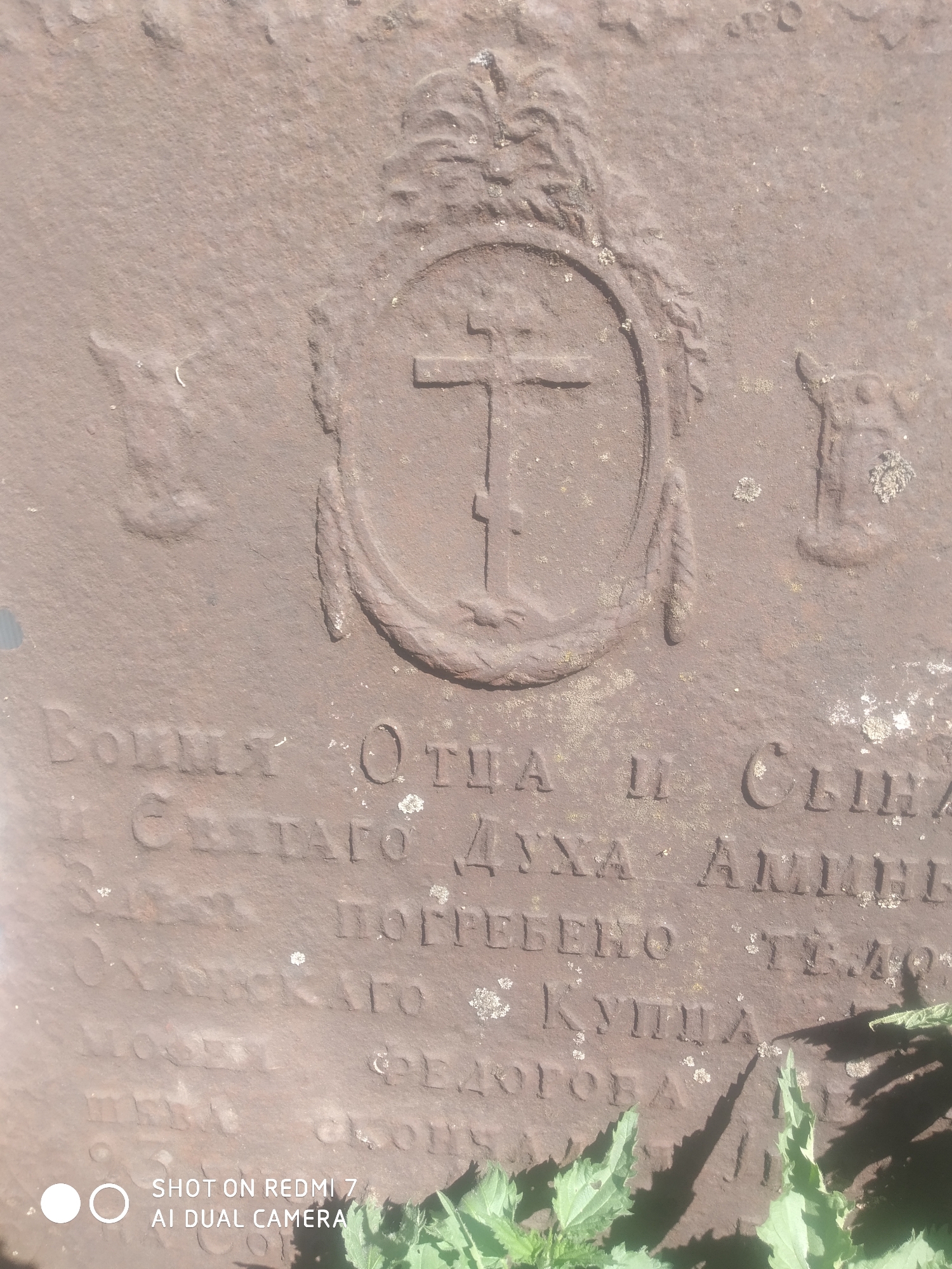The image depicts a weathered, possibly stone, grave marker or plaque. An ornate, circular emblem containing a cross with a slanted lower bar is centrally placed at the top. Below the emblem, inscription in Cyrillic script is visible. The inscription starts with the words "ВОИмя ОТЦА И СЫНА И СВЕТАГО ДУХА АМИНЬ" - "In the name of the Father and of the Son and of the Holy Spirit Amen." The inscription continues "ПОГРЕБЕНО ТЕЛО" - "Body buried", with part of an individual's name appearing to read "... ФЕДОГОВА". There is some vegetation covering the bottom of the plaque.
While the image itself does not provide specific location information, based on the text, which appears to reference a person from "Оханского" (Okhansk), it is reasonable to infer that the marker could be located in or near the city of Okhansk, Russia.