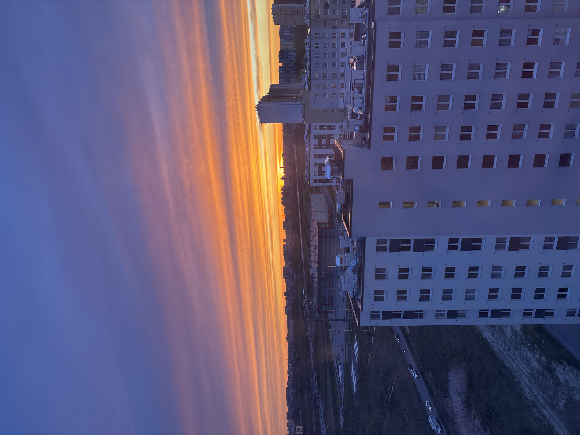 The image captures an urban scene at dusk, likely taken from a high vantage point. The sky is divided into two distinct color zones: a gradient of orange and yellow hues near the horizon, transitioning to a deep blue higher up. Linear cloud formations stretch horizontally across the sky, mirroring the city layout below.
To the right of the sky, a tall, modern apartment building with a uniform grid of windows dominates the frame. The building's facade is a pale color, possibly white or light gray, contrasting with the darkening sky. Below the building, a road runs parallel to the horizon, with cars visible along its length. Beyond the road, there is a strip of green space, possibly a park or a line of trees.
In the distance, the silhouette of other buildings can be seen along the horizon, creating a layered effect. The overall impression is that of a city landscape at the cusp of night, with the last light of the sun illuminating the sky and buildings.