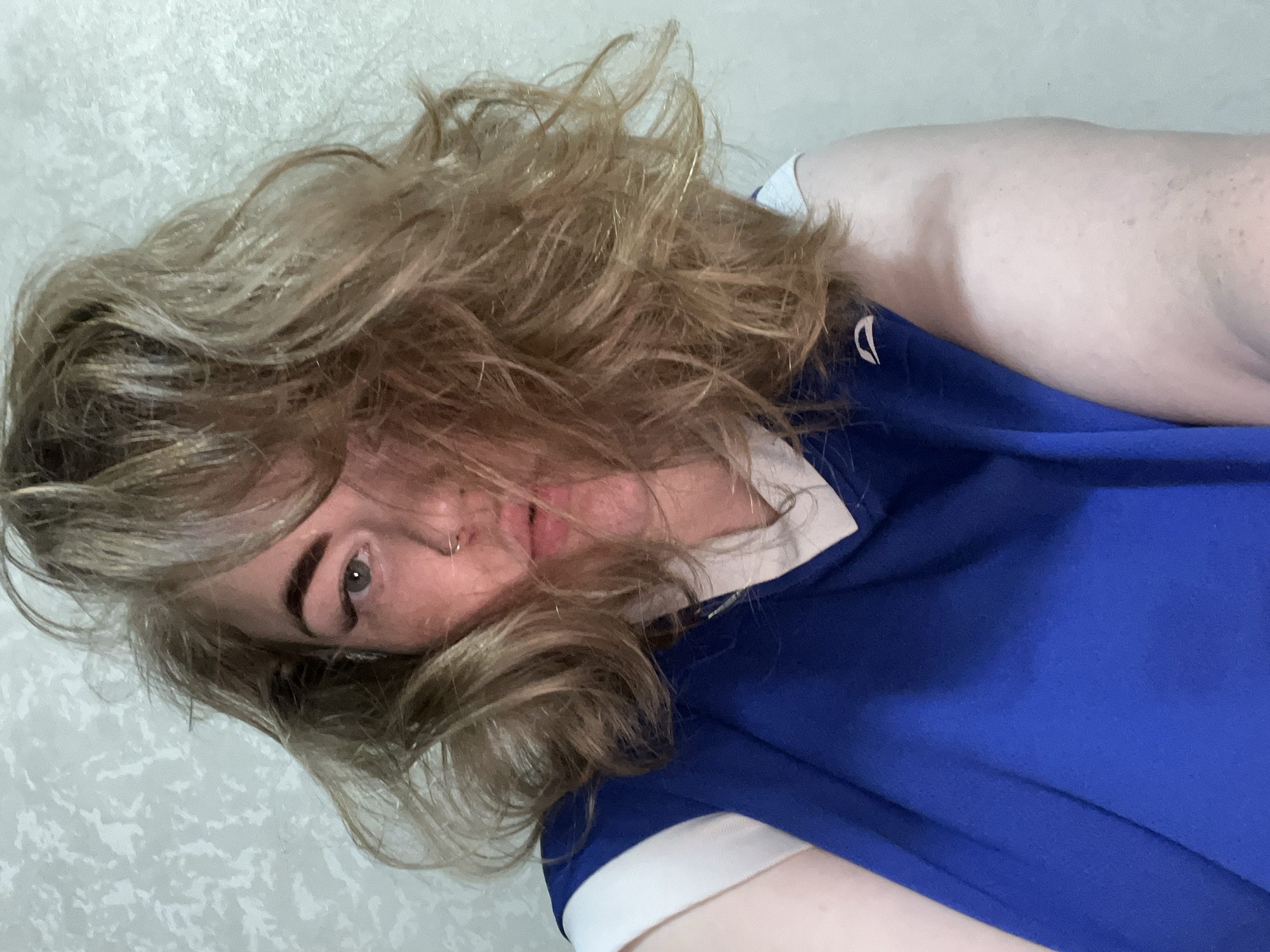 This close-up portrait features a person with their head tilted, or the image rotated ninety degrees clockwise, suggesting they might be lying down. The subject has voluminous, light brown to blonde, wavy or curly hair that largely frames and partially obscures their face. One eye, appearing green or hazel, is clearly visible, along with a portion of the nose, which features a silver hoop piercing on the left nostril. A hint of the mouth is also discernible. The person is wearing a bright royal blue shirt, which has a distinct white collar or trim, resembling a polo shirt or sports jersey. Fair-toned skin is visible on what appears to be an arm or shoulder. The background is a light, textured wall, likely off-white or light gray, indicating an indoor setting. There is no visible text in the image. The photo provides a close and intimate perspective, and the location is contextualized as Chaikovsky, Russia.