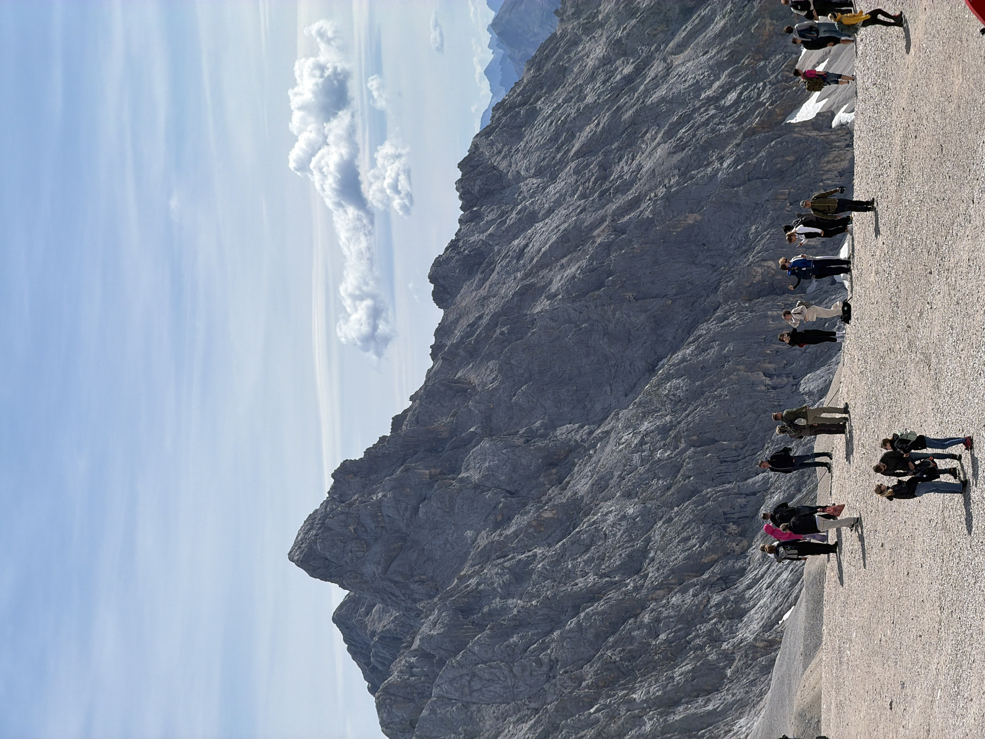 The image depicts a group of tourists standing on a gravelly plateau with a massive, rugged mountain range dominating the background. The sky is a light blue with scattered white clouds. The people, dressed in casual outdoor clothing, are scattered across the plateau, some in small groups and others individually. Some appear to be taking pictures or looking at the scenery. The mountain face is steep and rocky, with visible striations and shadows suggesting its immense scale. Patches of snow are visible on the slopes in the distance. The scene suggests a high-altitude tourist location, likely a scenic viewpoint. The location is identified as the Zugspitze, a mountain on the border between Germany and Austria, specifically near the summit area which is a popular tourist destination. Wildermieming is a municipality in the district of Innsbruck-Land, Tyrol, Austria, but the Zugspitze is not located within its administrative boundaries, though it is geographically close.