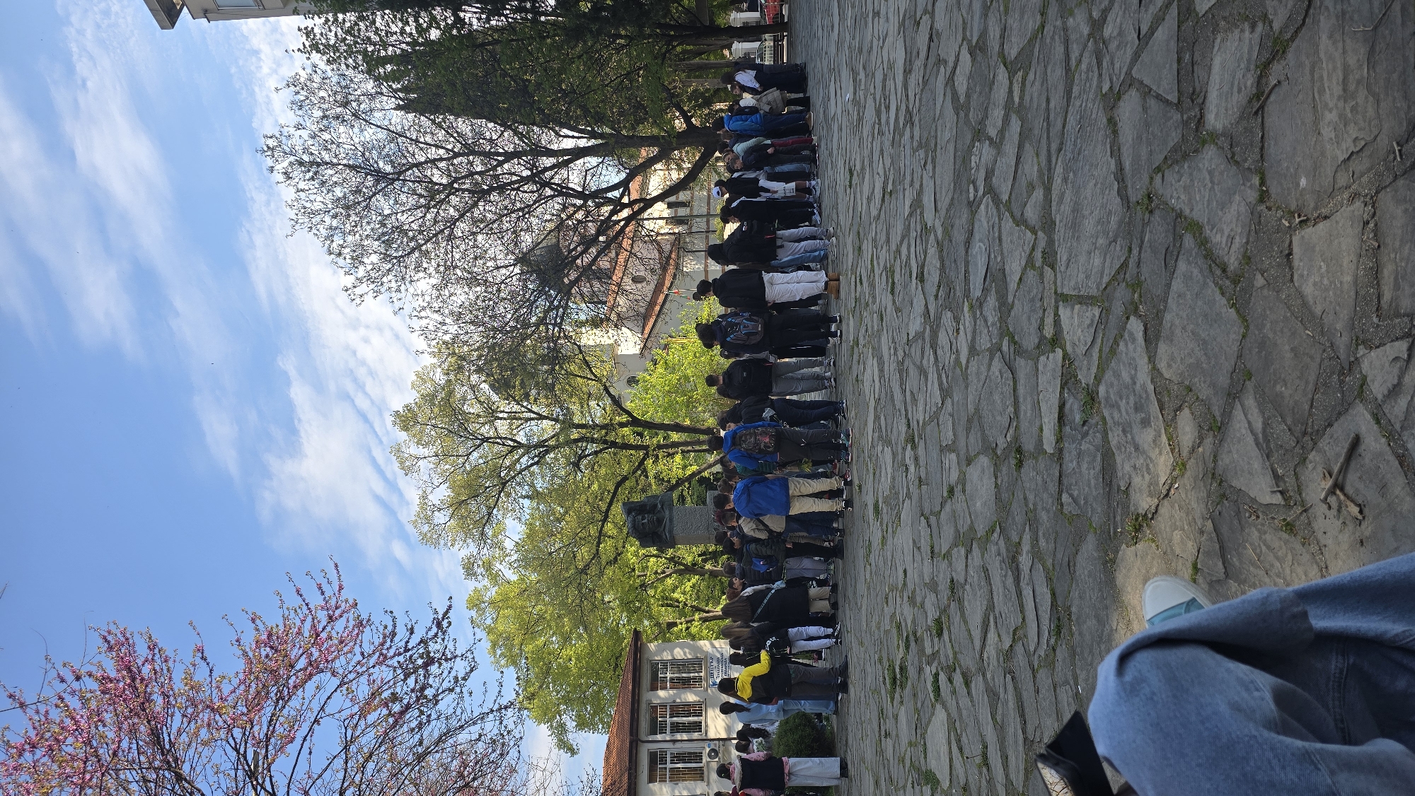 A long, narrow, full shot, oriented vertically, depicts a large group of people standing in a line in front of what appears to be a monument or statue, likely located in a public square or park in Stara Zagora, Bulgaria, based on architectural style and visible signage. The queue of people, mostly dressed in casual attire including jackets and backpacks, stretches away from the viewer. A cobblestone-style pavement is evident. In the background, there are trees with fresh spring foliage and buildings with red-tiled roofs. A bright blue sky with wispy clouds is above. A blossoming tree with pink flowers is visible on the left. In the bottom right corner, part of a person's leg, wearing jeans and a sneaker, is visible.