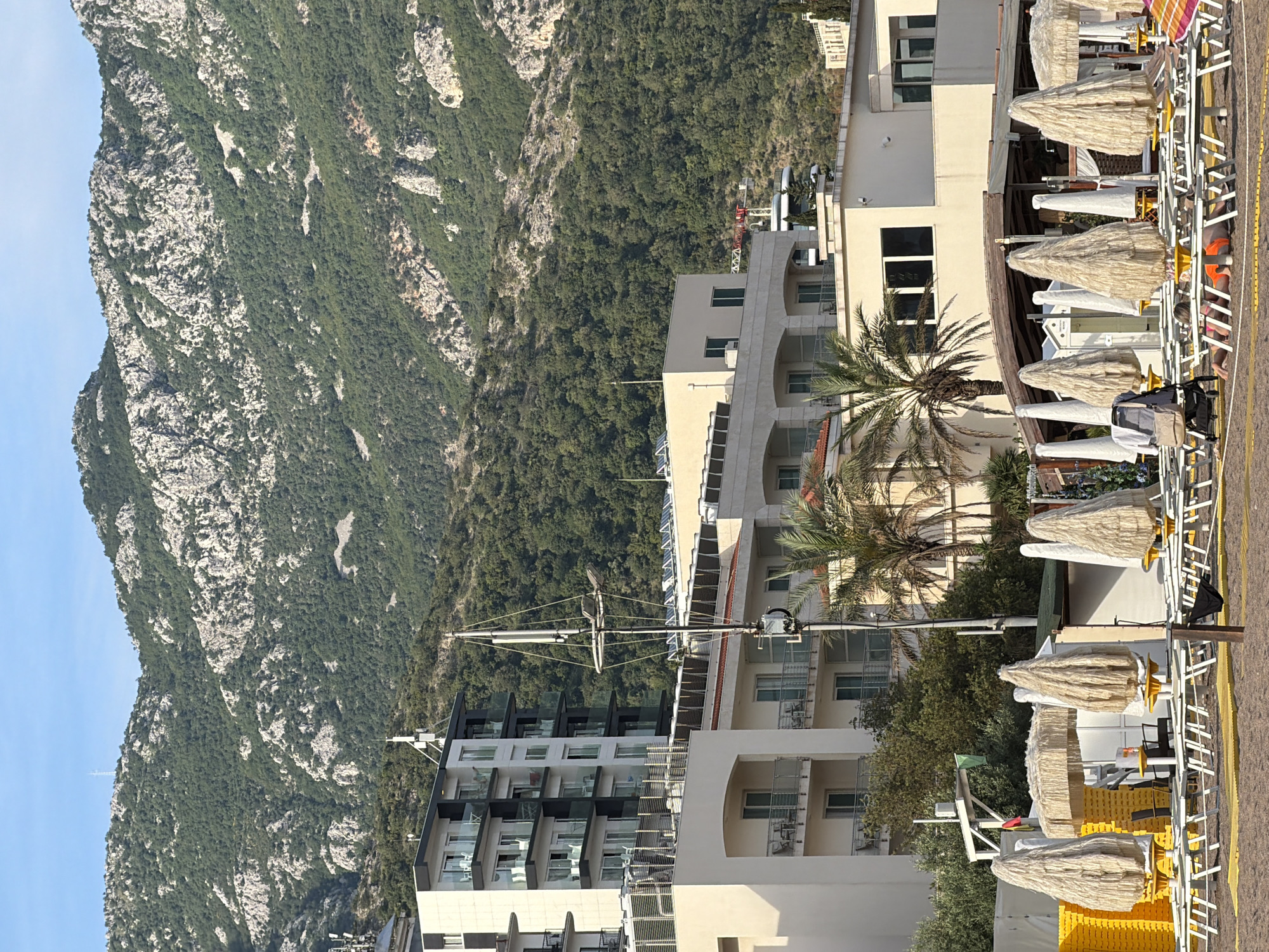This image captures a panoramic view of Boreti, Montenegro, featuring a multi-layered scene from a sandy beach to towering mountains. The background is dominated by large, verdant mountains, heavily forested with numerous grey rock outcrops and peaks. A tall, slender white telecommunications tower is visible on the highest distant peak under a clear blue sky.
In the mid-ground, a series of modern, multi-story buildings are visible, likely hotels or apartments. These structures vary in design, including white and dark grey facades, multiple balconies with glass railings, and some rooftops equipped with solar panels. Several cream-colored buildings with arched balcony structures and red-tiled roofs are also interspersed among the taller, more contemporary buildings. Numerous communication antennas and support poles are mounted on various rooftops and extend upwards from the mid-ground. Palm trees are visible between and in front of these buildings.
The foreground presents a sandy beach area. Numerous beach loungers, predominantly white, are arranged in rows, many resting on yellow mat sections. A prominent feature is the collection of closed beach umbrellas, which are covered in a shaggy, light-colored, possibly straw or thatch-like material, giving them a distinct textured appearance. One multi-colored striped umbrella is partially visible on the far right. A small, open-air beach bar or structure with a wooden roof is located behind some of the loungers. A stack of yellow beach mats is visible on the left side of the beach area, adjacent to a white frame structure displaying a small green flag. A grey baby stroller is positioned next to one of the loungers. One individual is lying face down on a lounger, wearing an orange swimsuit. The scene is illuminated by natural daylight, indicating clear weather.
