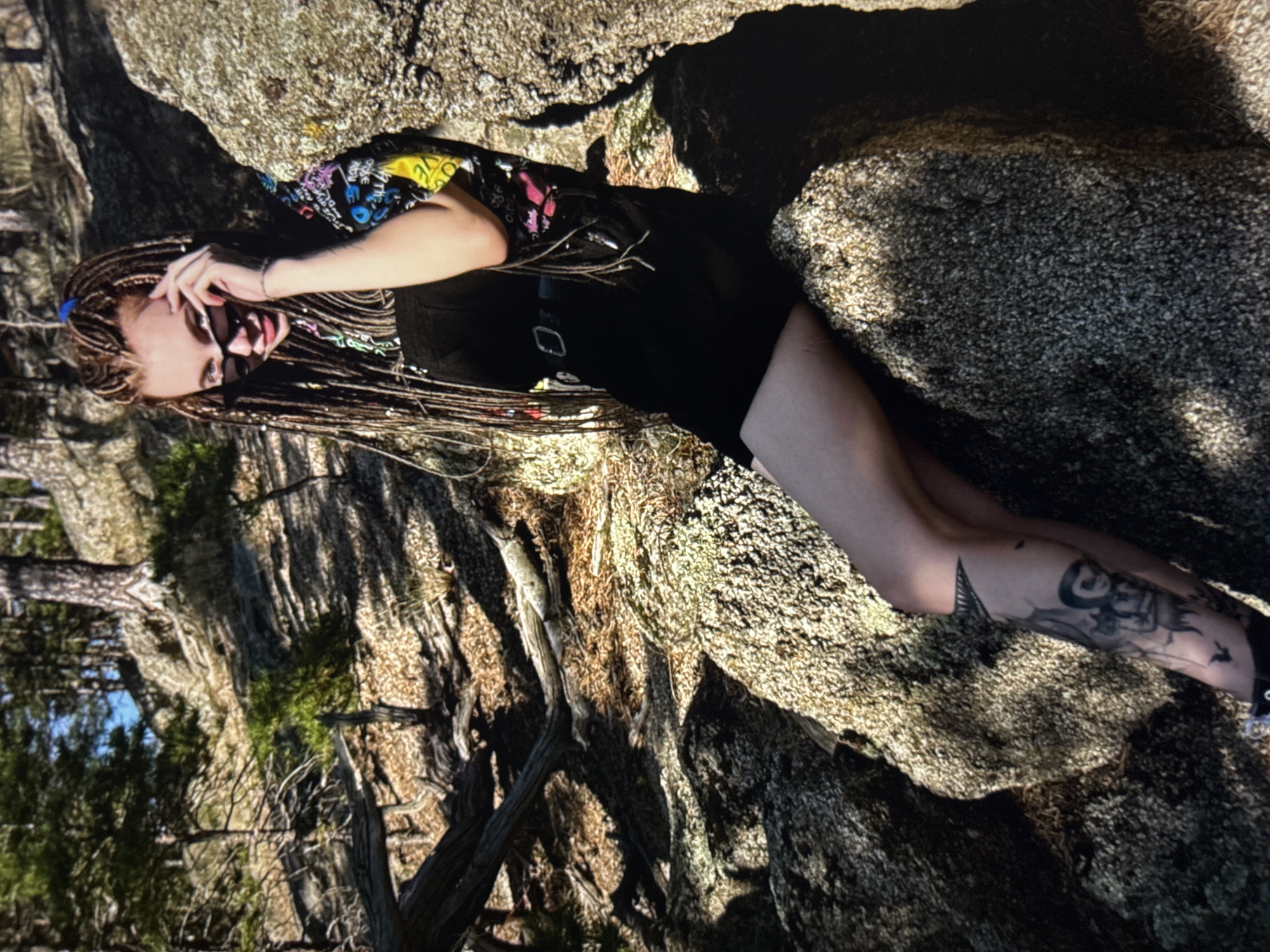 A young woman with blonde dreadlocks and sunglasses poses outdoors, leaning against a large rock in a wooded area. She is wearing a black dress with a belt and a t-shirt with a colorful graphic print. Her left leg is visible and shows a tattoo of a figure with wings and a bird. The background consists of trees, rocks, and dry foliage, suggesting a natural, possibly mountainous or hilly, environment. The lighting indicates it is daytime, with sunlight casting shadows. There is no visual information in the image to identify the location as Omsk, Russia.