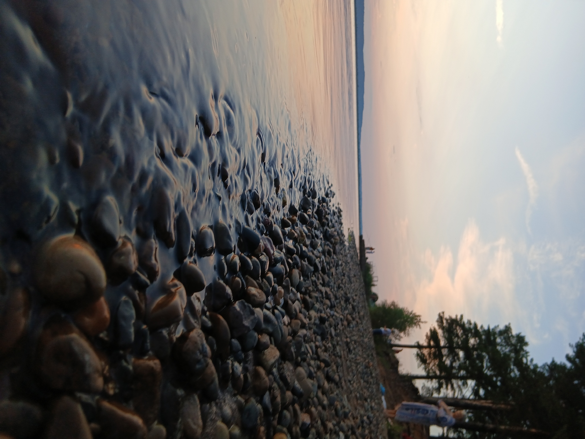 This is a low-angle, close-up shot of a rocky shoreline next to a body of water, likely a lake or river, during a sunset. The foreground is dominated by smooth, wet, grey and brown pebbles. The water's surface has ripples and reflects the soft, pastel colors of the sky, with hues of pink, orange, and blue. In the mid-ground, the shoreline continues with more pebbles and a few scattered green trees. In the background, on the higher bank, several people are visible. One person, a woman, stands out, wearing a light-colored robe or dress, looking towards the water. Further back, more figures are scattered, some appearing to be walking or standing. The sky is mostly clear with a few wispy clouds, and a distant, blurry mountain range can be seen on the horizon. The overall atmosphere is serene and tranquil.