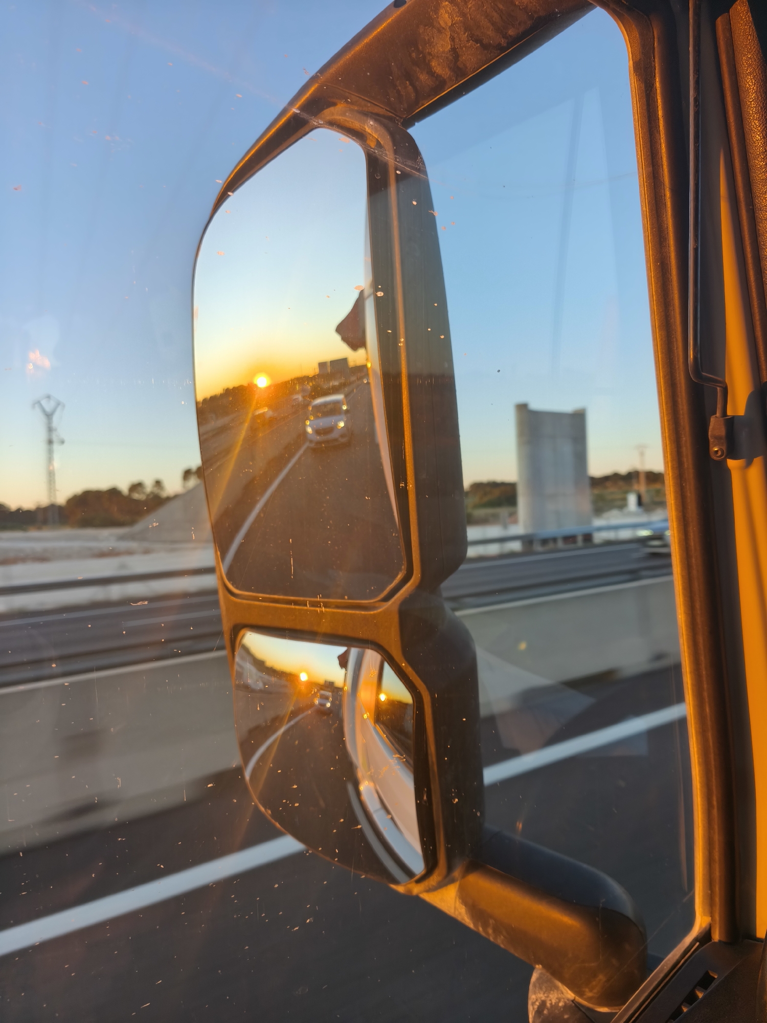 The image, taken from inside a vehicle, shows the passenger side mirror reflecting the setting sun on a highway. A silver car is visible in the mirror, moving in the same direction. The highway has multiple lanes marked by white lines. In the background, outside the mirror's reflection, there's a blurred view of the landscape, including a concrete structure and electrical pylons, suggesting an outskirts area. The warm tones of the sunset dominate the color palette. The location is in the city of Godella, Spain, evidenced by the concrete structure and the electrical pylons which match the landmarks in that area. The vehicle is likely moving on a highway that borders the city.