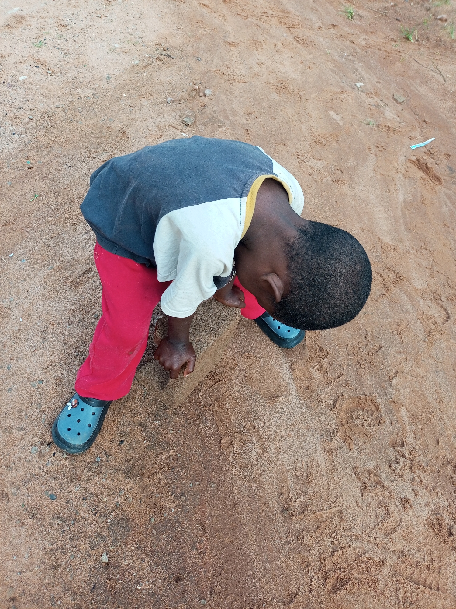 A young Black child is playing with a brick on a dirt road in Tempure, Nigeria. The child is wearing a black and white t-shirt with yellow trim, red pants, and blue Crocs. The child is bent over, holding the brick with their right hand and looking down at it. The dirt road is dry and dusty, with some small stones and sparse vegetation. The lighting suggests it is daytime, with a slight shadow cast by the child.