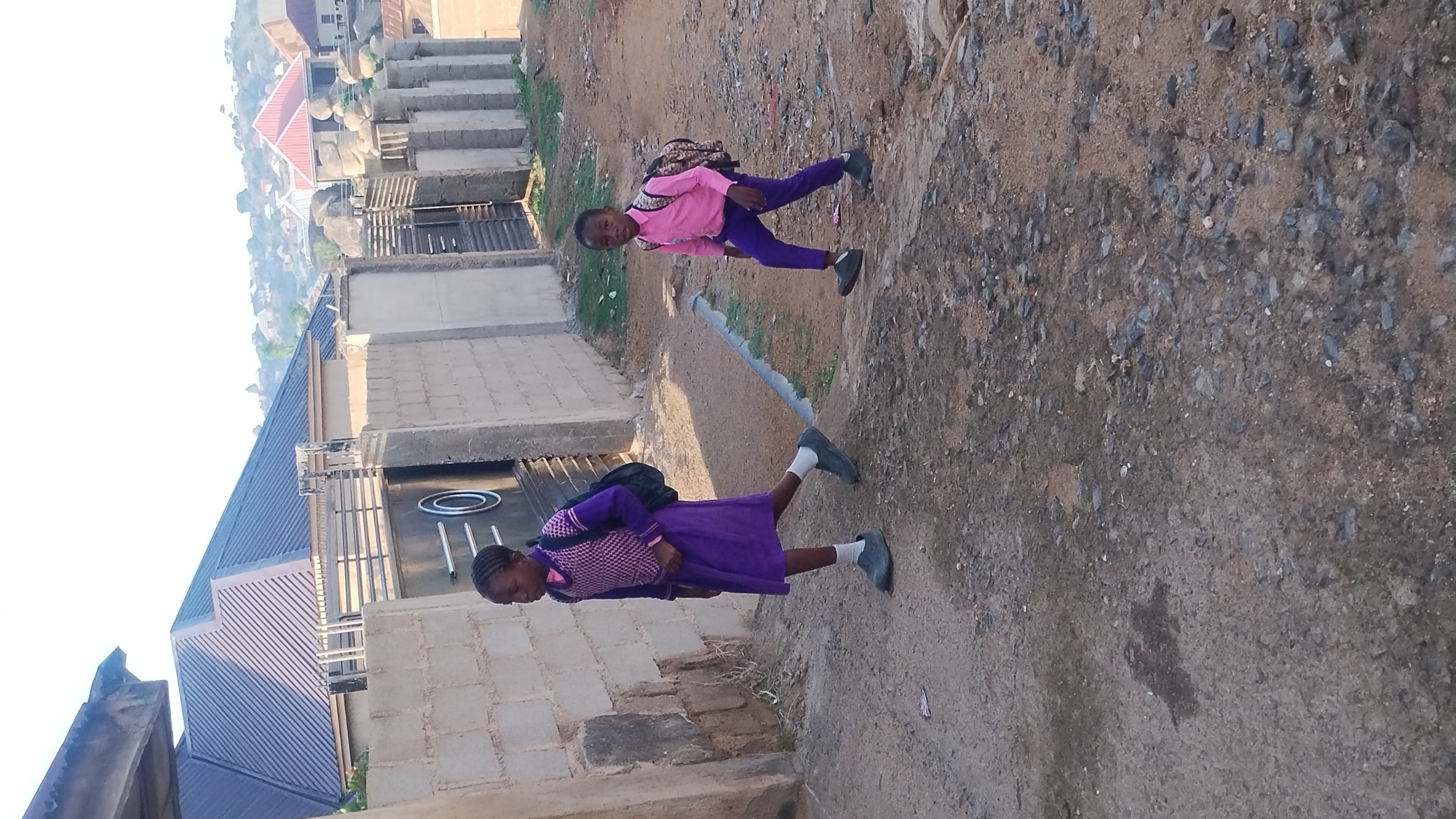 The image displays two young girls standing on an unpaved dirt and gravel path in a residential area, under bright daylight conditions. The photograph is oriented vertically, causing the subjects and environment to appear tilted to the right.
In the foreground, on the left side of the path, a girl wears a purple dress or tunic over a light-colored, possibly checkered, patterned top. She has a dark backpack on her shoulders, white socks, and dark shoes. Her head is slightly bowed, suggesting she might be in motion or looking down.
Further along the path, positioned towards the center-right, stands another girl. She is dressed in a bright pink long-sleeved shirt, purple pants, and dark shoes, also carrying a backpack. This girl is looking directly at the camera with a neutral expression.
The background features several buildings constructed with cinder blocks, many of which have dark metal gates and corrugated metal roofs. Beyond these structures, a distant landscape of hazy hills or mountains is visible under a clear, bright sky, indicating daytime. The ground consists of loose dirt mixed with numerous small rocks and pebbles. There is no visible text or explicit branding in the image. The scene conveys an outdoor, everyday setting, possibly in a developing or rural residential community.