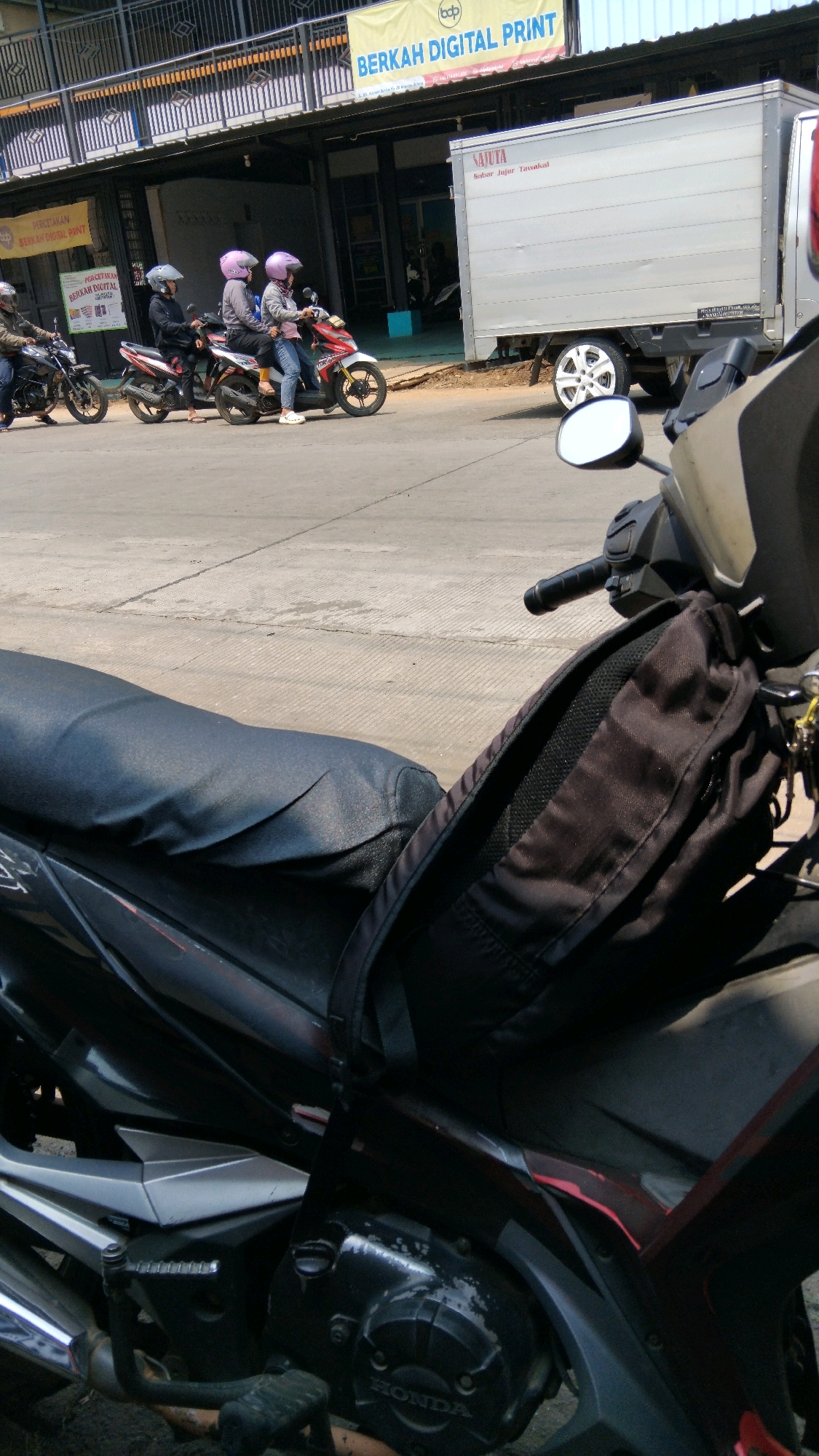 The image shows a street scene in Batang, Indonesia, with a focus on a motorcycle in the foreground. A black backpack is hanging from the handlebars of the motorcycle. In the background, three people are riding a motorcycle. They are all wearing helmets, and the two passengers in the back appear to be women. Behind them, a white box truck is parked. A yellow banner with Indonesian text hangs from a building. The street is paved, and there are some trees and foliage visible. The overall impression is of a busy street in a city.