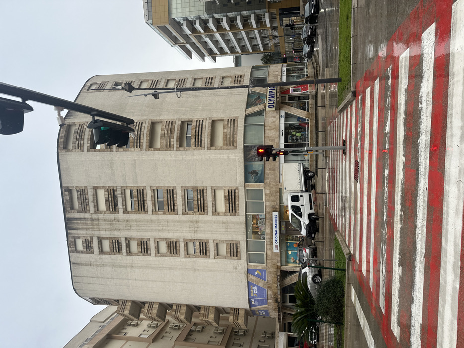 This image depicts an urban street scene in Budva, Montenegro, specifically near Mediteranska street. The weather is overcast, and the street surface is wet, indicating recent rain.
The central subject is a nine-story, light-colored commercial and residential building with a curved facade and numerous horizontal rows of windows, many featuring external roller blinds. Vertical streaks are visible on the building's exterior. The ground floor houses retail businesses, including "ORIGINAL MARINES" and "OLYMPIC" stores, identifiable by their signage, alongside a sign for "PRVA BANKA CRNE GORE."
In the foreground, a distinctive pedestrian crosswalk with alternating red and white stripes extends across the wet asphalt. A traffic light positioned within the crosswalk displays a red signal, while an overhead traffic light shows green. A white panel van with a canvas-covered flatbed is stopped or parked in the street in front of the "Original Marines" store. Multiple dark-colored passenger vehicles are visible parked or moving further down the street to the right. A tall, dark street light pole stands to the right of the crosswalk. A small green median with a palm tree and shrubs is situated to the left of the crosswalk. A white security camera is mounted on a pole visible near the top left of the main building. No individuals are prominently featured or engaged in specific actions within the frame.