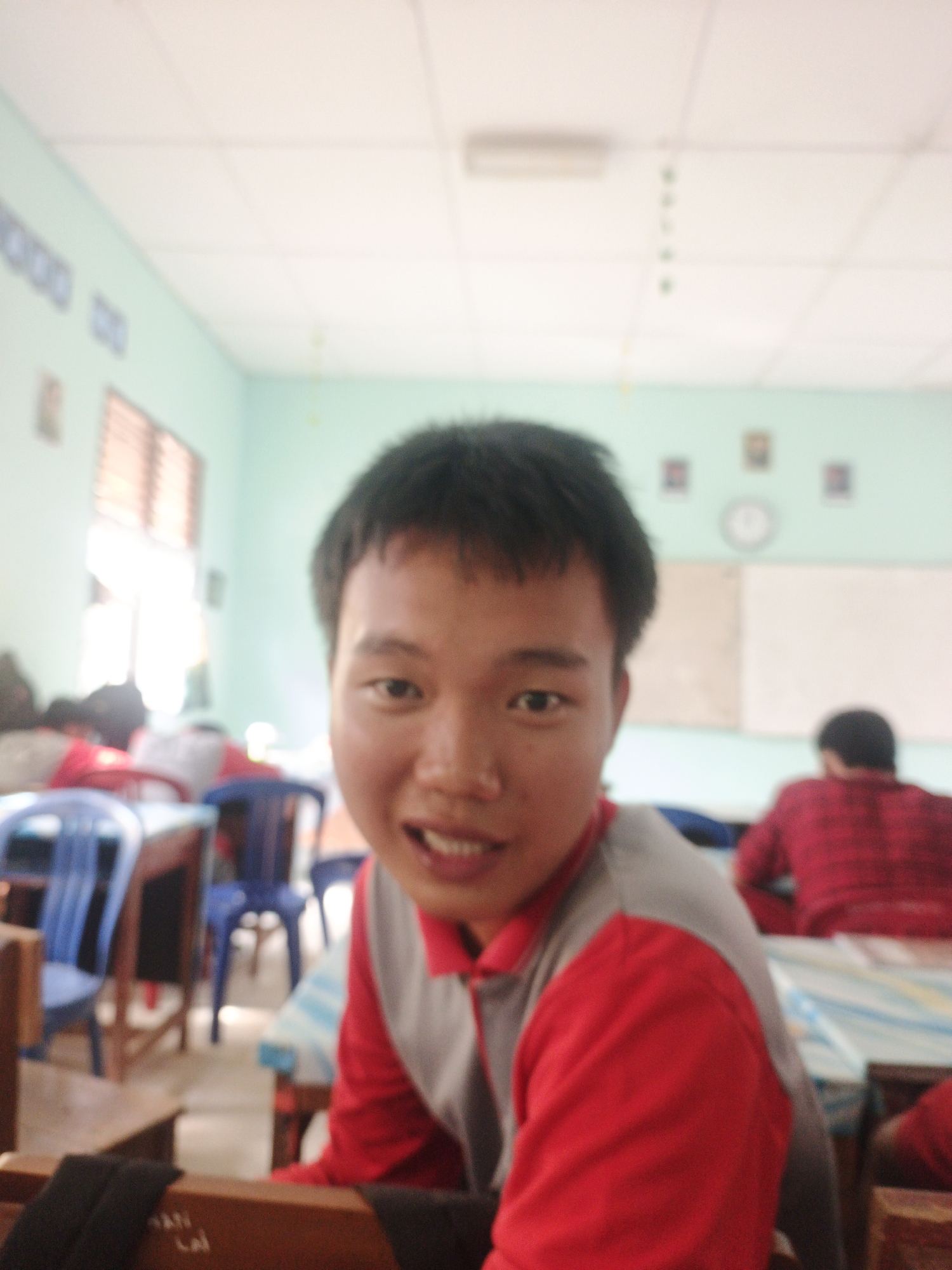A slightly blurry, eye-level medium shot captures a young East Asian male in the foreground, facing forward with a slight smile, and looking at the camera. He has dark, short hair, and is wearing a red and grey shirt. The background is a classroom setting. The walls are painted a light green, and a white ceiling has fluorescent lighting. Several framed pictures and a clock are visible on the wall. Desks and blue chairs are arranged in rows. In the background, another person wearing a red shirt is visible from the back. The overall scene suggests a casual indoor environment, likely a school. The prompt mentions Jepara, Indonesia, but there are no visual cues in the image to confirm this specific location.