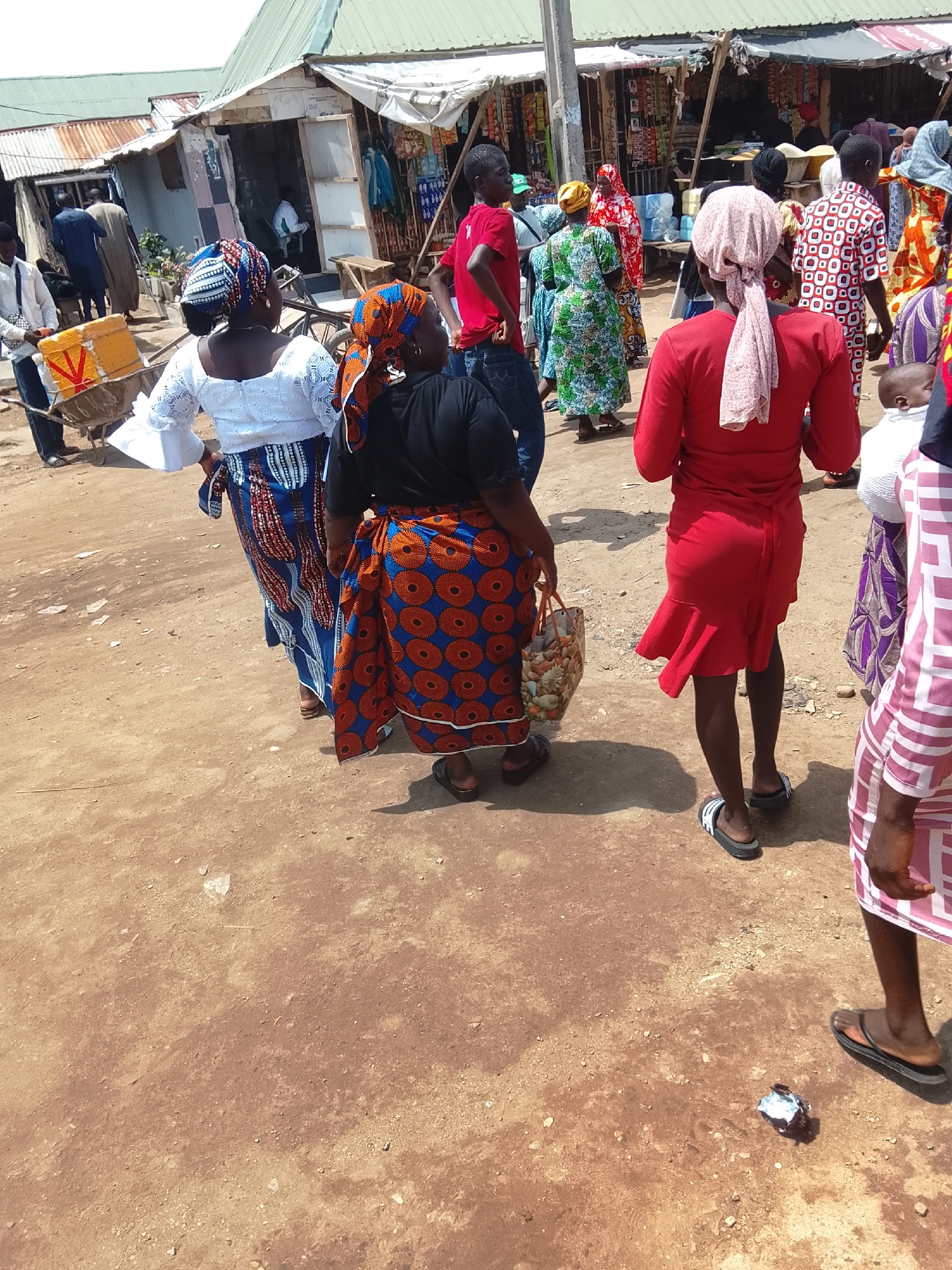 This is a medium shot, eye-level, outdoor photograph taken in a marketplace setting in Abuja, Nigeria. The scene depicts a bustling environment with several people, predominantly women, walking and interacting. The ground is a dusty, light brown dirt surface, indicative of a market or unpaved area.
In the foreground, a woman from the back, wearing a vibrant red dress and a pink headscarf, is walking away from the camera. She is wearing black flip-flops. To her left, another woman, also seen from the back, wears a black top and a brightly patterned skirt with orange circles on a blue background. She is carrying a woven bag filled with items. She is wearing flip-flops.
Further back, a woman in a white top with flared sleeves and a patterned skirt in blues and reds is visible. She is standing next to a yellow box with red markings, which appears to be a container, placed on a wheelbarrow.
Other individuals are present in the background, including a man in a red t-shirt on the far left, and several people in colorful clothing, suggesting they are shoppers or vendors. The background shows market stalls with various goods displayed, and some rudimentary structures with metal and corrugated iron roofs, typical of a Nigerian open-air market. The overall atmosphere appears to be one of everyday commerce and activity.