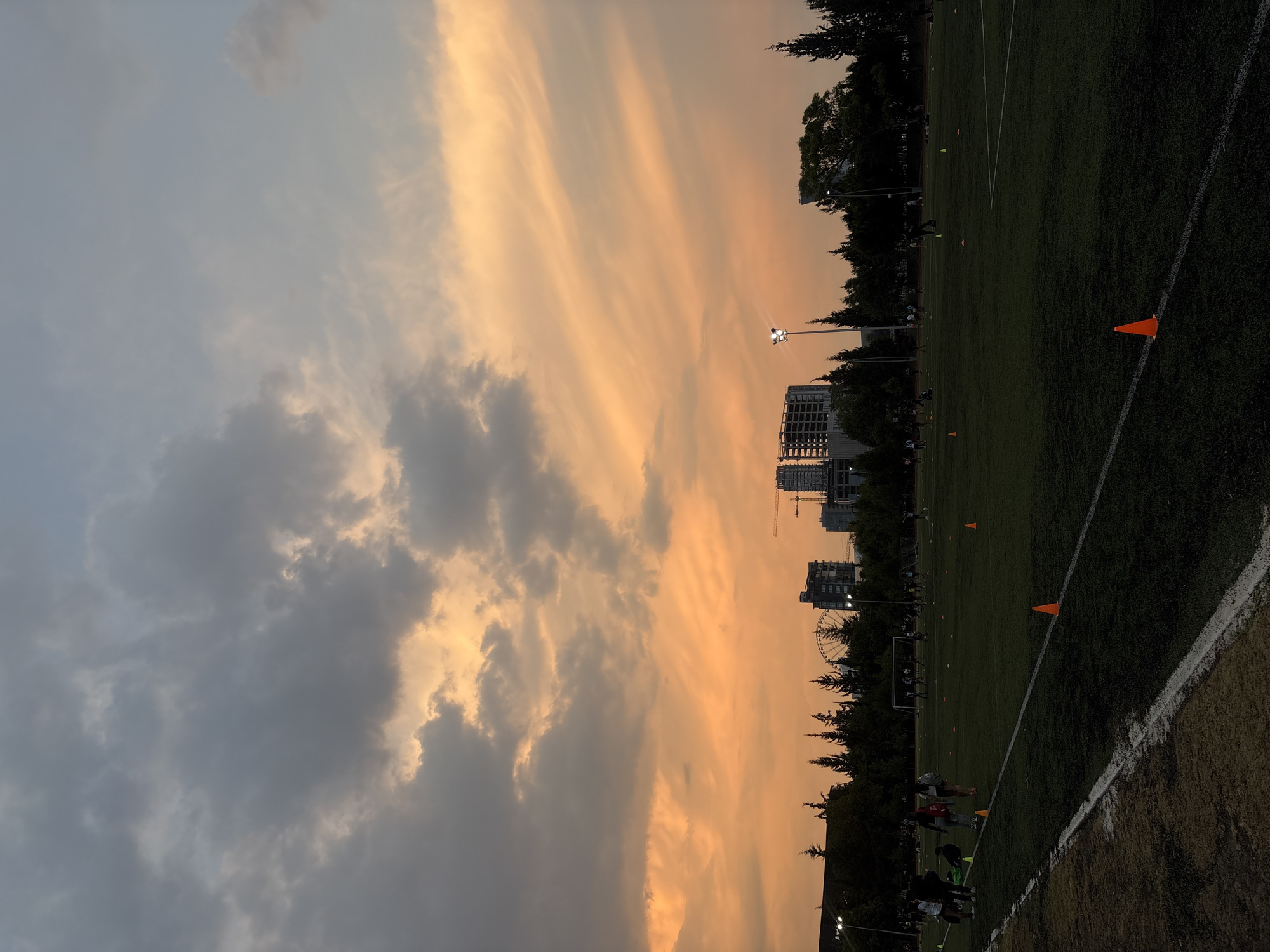 Here is a forensic description of the image.
**Scene**: The image appears to show a sunset over a sports field in the city of Puebla, Mexico.
**Sky**: The sky is a mix of colors. One side has darker, grey clouds, while the other side is illuminated with orange and yellow hues.
**Buildings/Objects**: In the middle ground, some buildings and a ferris wheel are partially visible. A construction crane is also noticeable in the background.
**Sports Field**: A green sports field dominates the foreground. Orange cones are placed at intervals along the field.
**People**: Although difficult to discern in detail, there are indistinct figures of people on the field, possibly engaged in some kind of activity or training.
**Overall Impression**: The image conveys a scene with a juxtaposition of natural elements (sunset and clouds) and urban elements (buildings, a ferris wheel, and a sports field).