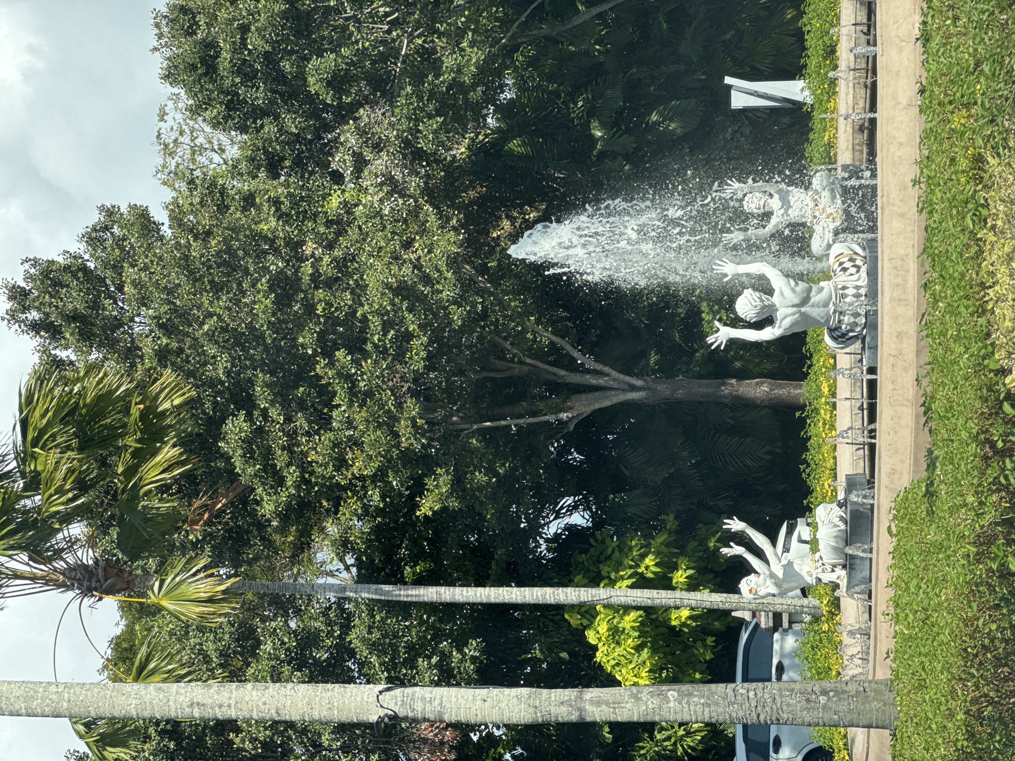 This image depicts a scenic view in Kuta Selatan, Indonesia, featuring a water fountain with three sculptures. The sculptures, made of a white material, are arranged in a semi-circular formation around a central fountain that shoots a powerful stream of water upwards. Two of the sculptures, positioned towards the back, are depicted with their arms raised, seemingly interacting with the water. The third sculpture, to the left and slightly forward, is seated with its arms extended and raised. Surrounding the fountain area are lush green trees and palm trees, creating a tropical ambiance. A white car is partially visible in the background, parked to the left of the fountain. The foreground is dominated by manicured green bushes. The sky above is overcast with white clouds.