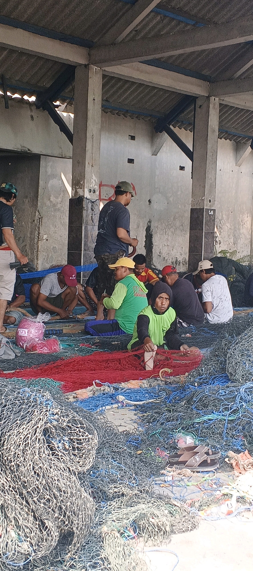 In Blimbing, Indonesia, a group of men are gathered under a corrugated metal roof supported by concrete pillars. The ground is covered with a large amount of fishing nets in various colors, including red, blue, and grey. Several men are seated on or around the nets, some appearing to be mending or sorting them. One man, wearing a green long-sleeved shirt, is actively working with a red net. Another man in a dark shirt and camouflage pants stands, holding a red circular object. A stack of pink netting is visible in the foreground. The environment appears to be an open-air structure, possibly a storage area or workshop related to fishing activities.