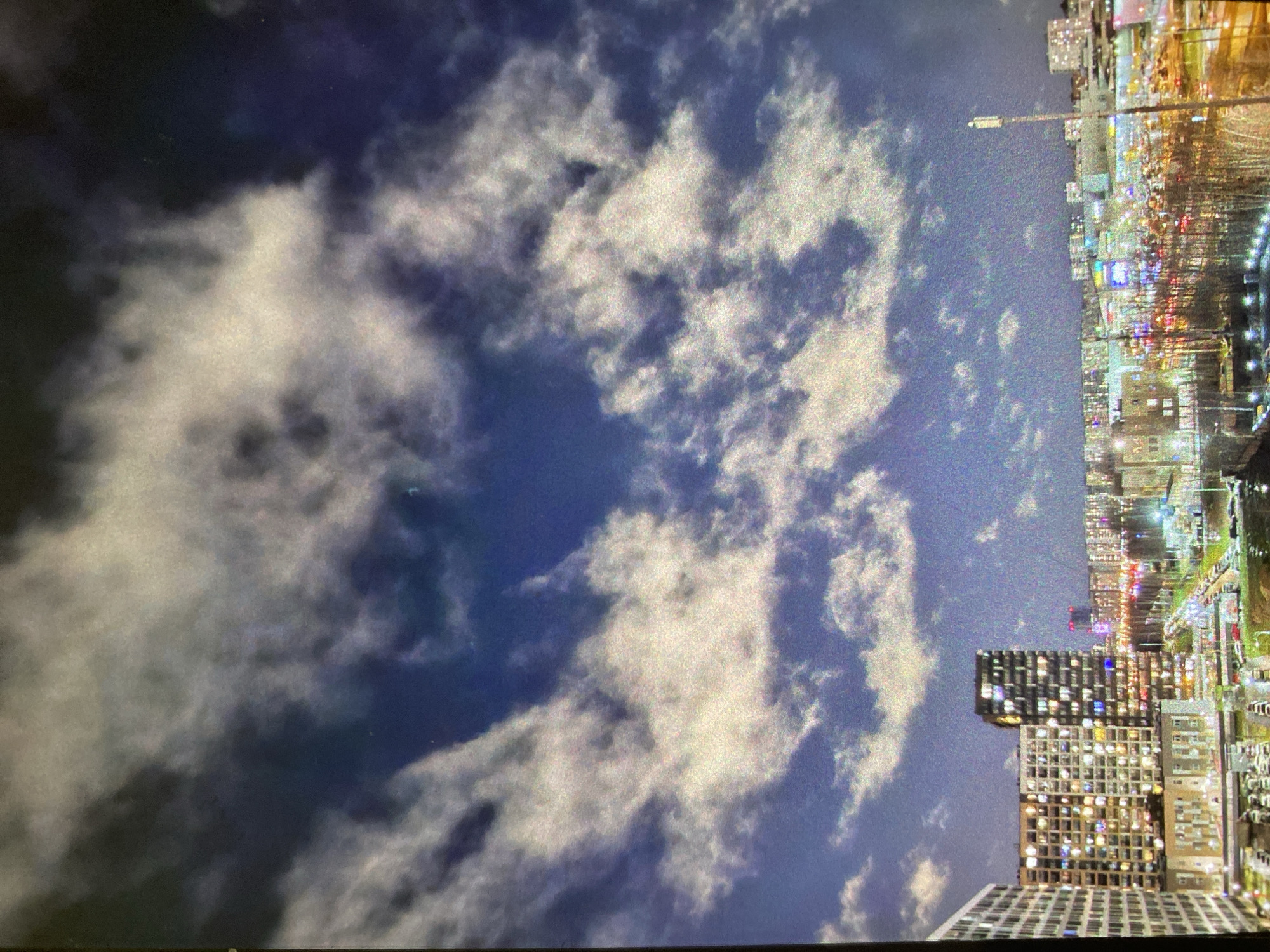 The image, oriented vertically, captures a striking night or dusk scene of a cloudy sky above a bustling cityscape. The sky is a deep blue, almost black in places, and is extensively covered by thick, textured white and grey clouds. These clouds appear to be illuminated from below by city lights, creating a dramatic contrast against the dark expanse.
The right side of the image (when rotated correctly) showcases a dense urban environment. Numerous tall buildings, likely residential or commercial, dominate the skyline, with countless windows brightly lit, indicating activity within. The artificial lights from these structures, along with streetlights and distant traffic lights, create a vibrant, warm glow across the city. A distinct streetlight is visible, extending upwards from the urban edge.
The time of day is clearly evening or night due to the dark sky and the reliance on artificial lighting. The weather is cloudy, but the clouds reflect the city's luminosity rather than completely obscuring the sky. The image exhibits a grainy or noisy quality, characteristic of low-light photography. There are no identifiable people or specific events occurring, nor is any visible text present. The scene depicts a lively metropolitan area, consistent with a major city like Saint Petersburg, Russia.