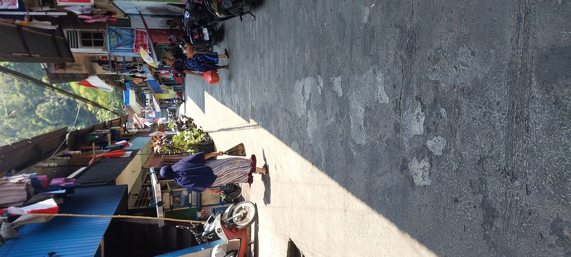 This is a vertical, eye-level, full shot of a narrow, sun-drenched alleyway in Jayapura, Indonesia. The alley is flanked by tightly packed, multi-story buildings with visible electrical wires crisscrossing overhead. The buildings exhibit a variety of materials, including blue metal siding, wood, and what appears to be corrugated metal roofing. Many windows and balconies are visible, some adorned with laundry hanging out to dry and Indonesian flags.
In the foreground, a woman wearing a dark blue hijab and a striped long dress walks away from the camera, carrying two shopping bags. To her left, a red and white scooter is parked. The alley surface is paved with concrete, showing signs of wear and tear with patches of lighter color. A distinct shadow of a building cuts across the alley, dividing it into a sunlit and shaded portion.
Further down the alley, another woman in a dark blue patterned dress walks towards the camera, carrying a red plastic bag. Behind her, several motorcycles are parked along the side of the alley. In the distance, more buildings and trees are visible, suggesting a hillside or elevated terrain. The overall impression is one of a densely populated urban environment, likely a residential area in Jayapura, characterized by its close-knit architecture and daily life activities.