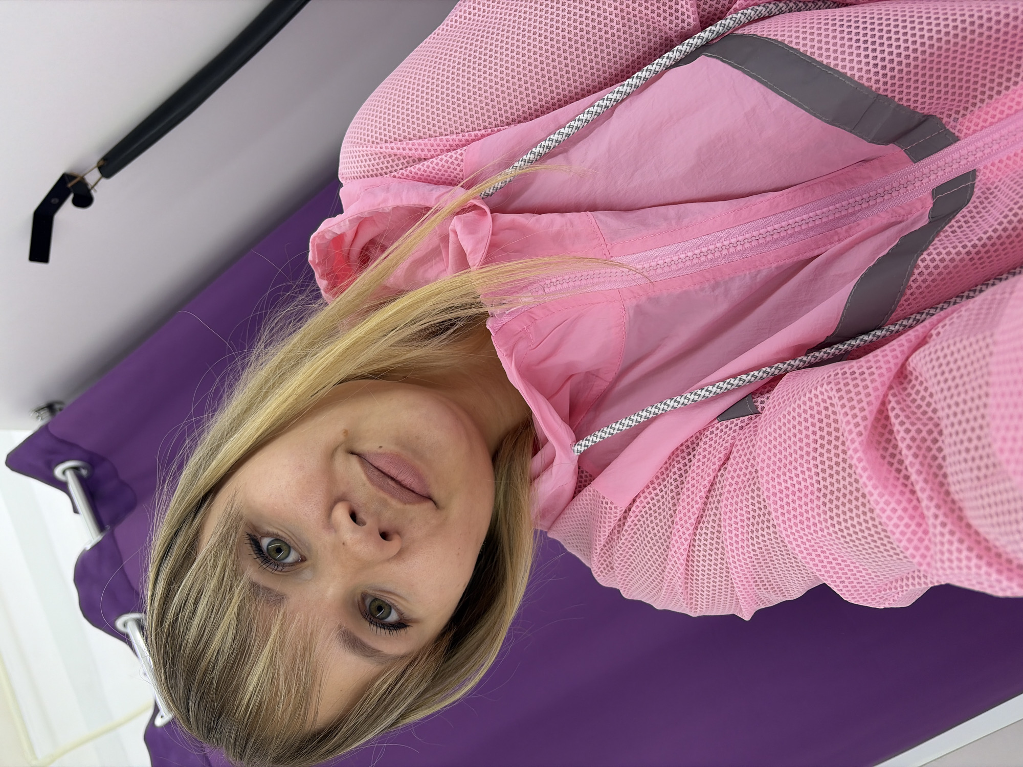 A young blonde woman with green eyes and fair skin is taking a selfie in what appears to be a fitting room. She is wearing a pink hooded jacket with a mesh overlay and reflective gray trim. The background consists of a purple curtain, a white wall, and a black pole attached to the ceiling. The woman is smiling slightly and her head is tilted. The image is taken from a low angle. There is no information available to determine the location within the city of Chaikovsky, Russia.