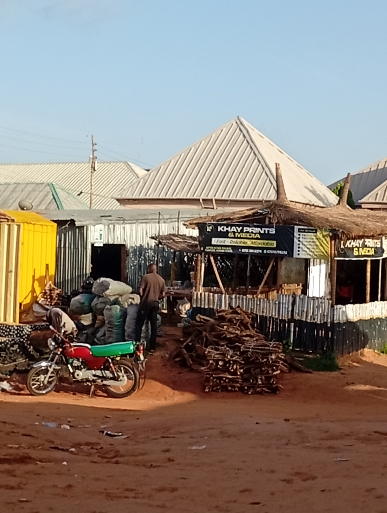 The image depicts an outdoor market scene in Tumfure, Nigeria. In the foreground, a red and green motorcycle is parked on a dirt path. To the left, a stack of firewood and a yellow shipping container are visible. Behind the motorcycle, a man in a brown shirt stands near a pile of large, dark sacks. Further back, a stall with a thatched roof displays a banner for "KHAY PRINTS & MEDIA." The stall is constructed with corrugated metal and wooden planks. Additional structures with corrugated metal roofs are visible in the background, suggesting a densely built area. The overall environment is dusty, with loose dirt and some scattered debris on the ground. The sky is clear and blue.