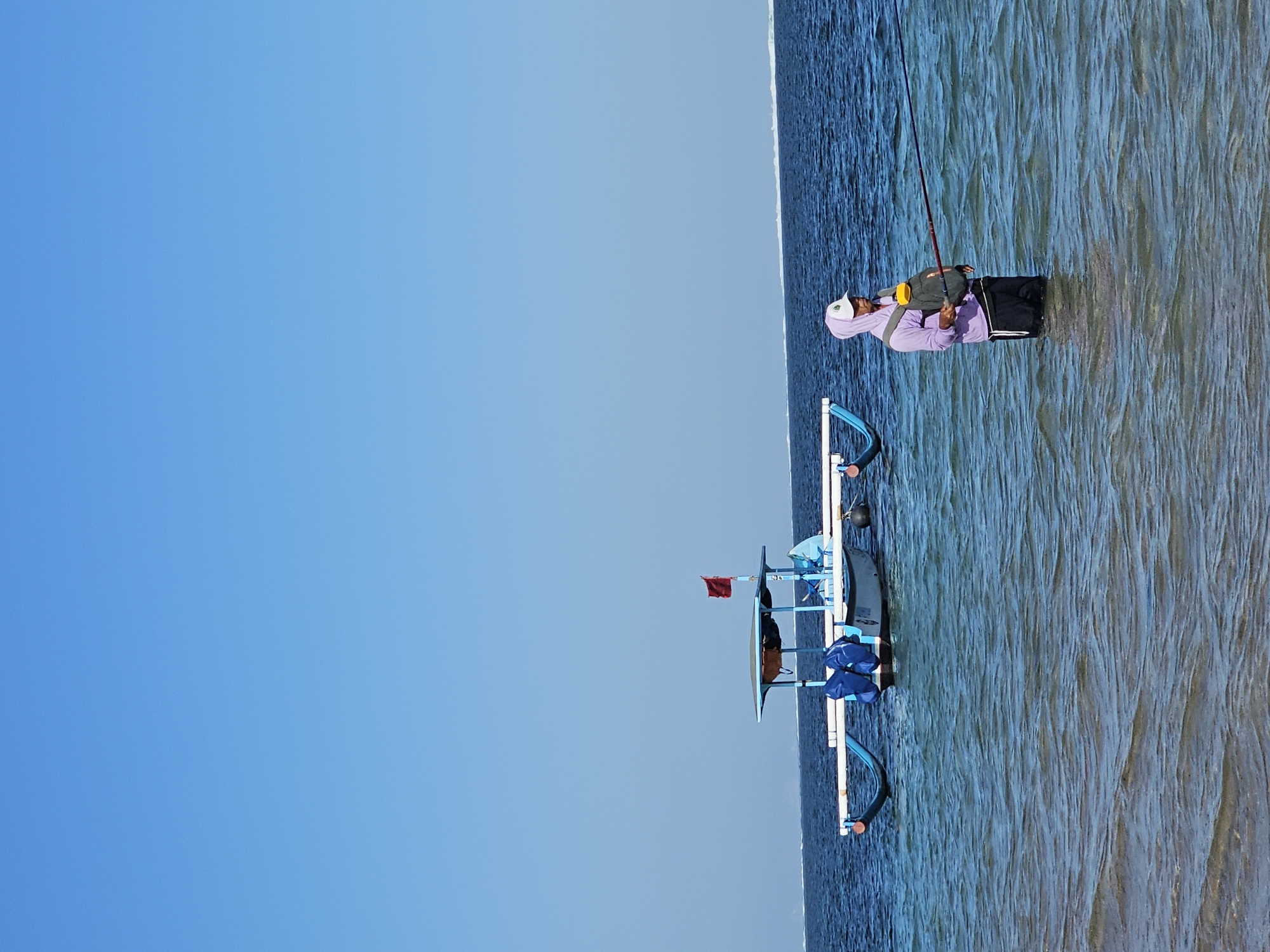 A person stands in shallow, clear ocean water, fishing. The person is wearing a purple hooded sweatshirt, a white cap, black shorts, and a green backpack. They are holding a fishing rod with the line in the water. To the left, a small blue and white boat with a red flag is anchored in the water. The horizon is visible in the distance with a thin white line of breaking waves. The sky is clear and blue. The scene appears to be a beach in Denpasar, Indonesia.