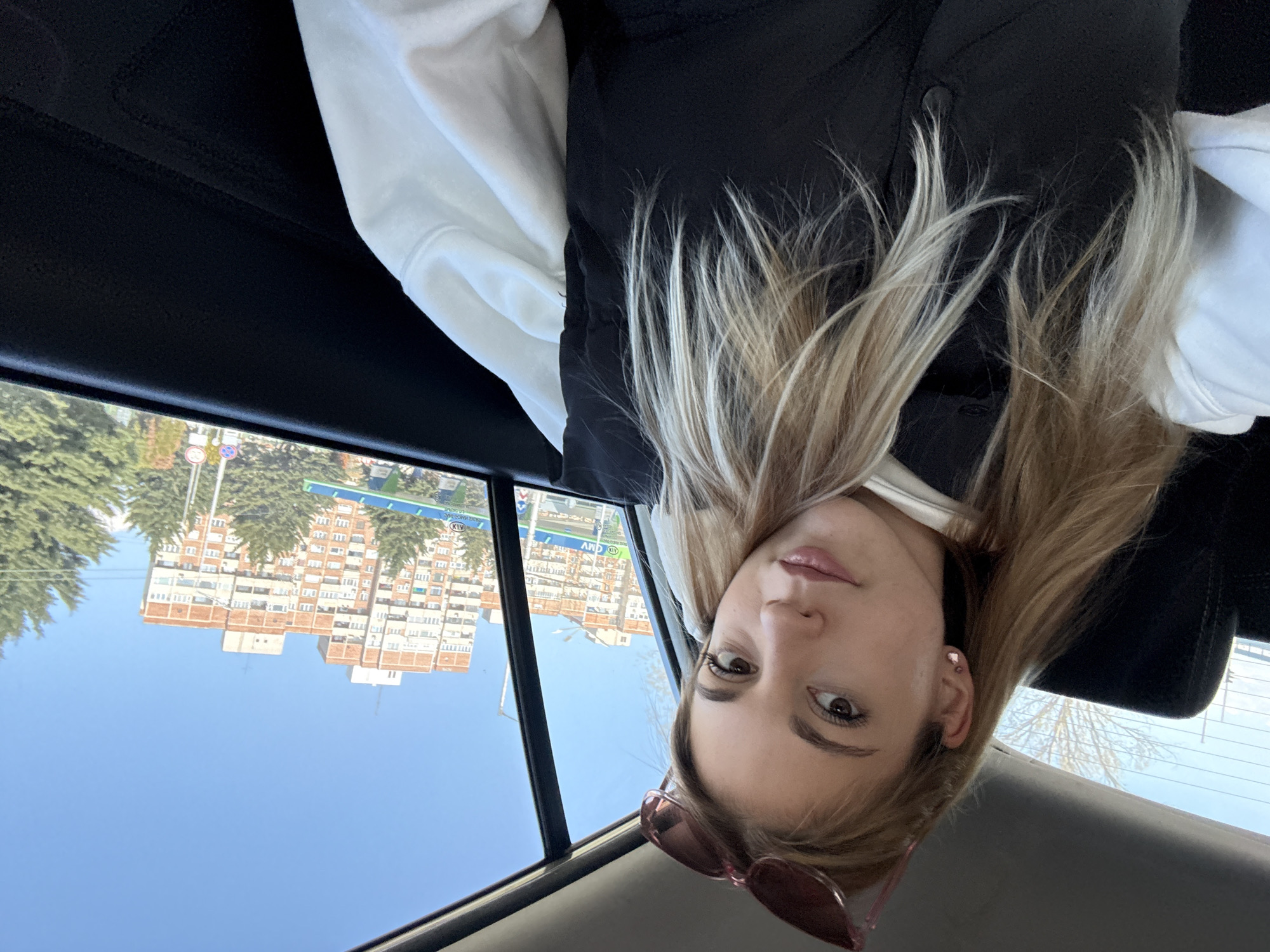 A woman is in the backseat of a car, wearing a white sweatshirt and a black vest. The image is taken from her perspective looking up towards the car's ceiling. The exterior background visible through the car window shows buildings and trees under a clear blue sky. The location appears to be Sofia, Bulgaria, based on the architecture and street signs visible in the background. The woman is wearing pink sunglasses and has light brown/blonde hair.