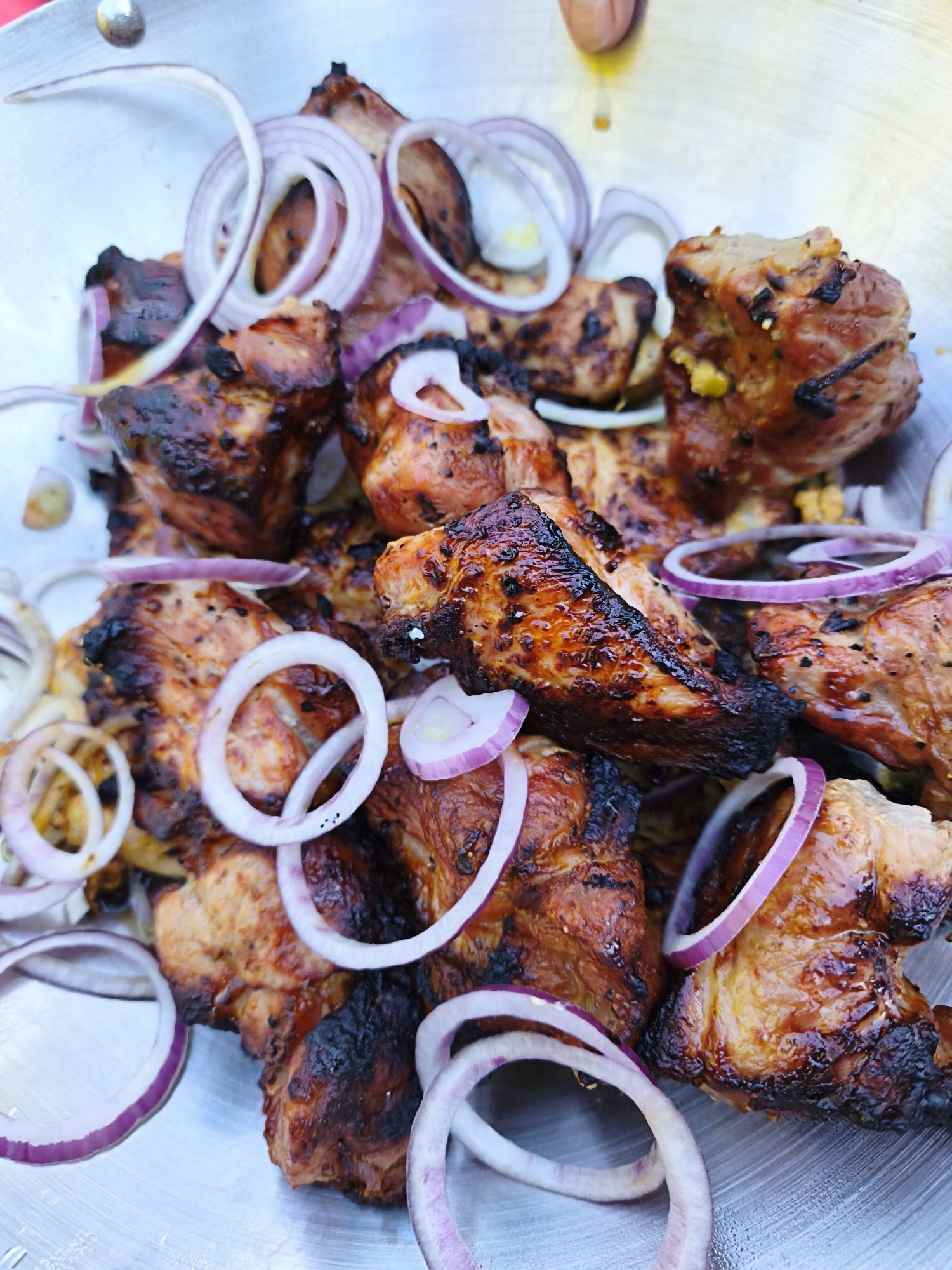 This is a close-up, overhead shot of grilled meat and red onion rings in a metal bowl. The meat is cut into bite-sized pieces and appears to be pork or chicken, with visible grill marks and some charring. The red onion rings are sliced thin and are scattered amongst the meat. The lighting is bright, casting some shadows. The image does not depict any identifiable landmarks or details that would allow for the determination of its location within Maardu, Estonia.