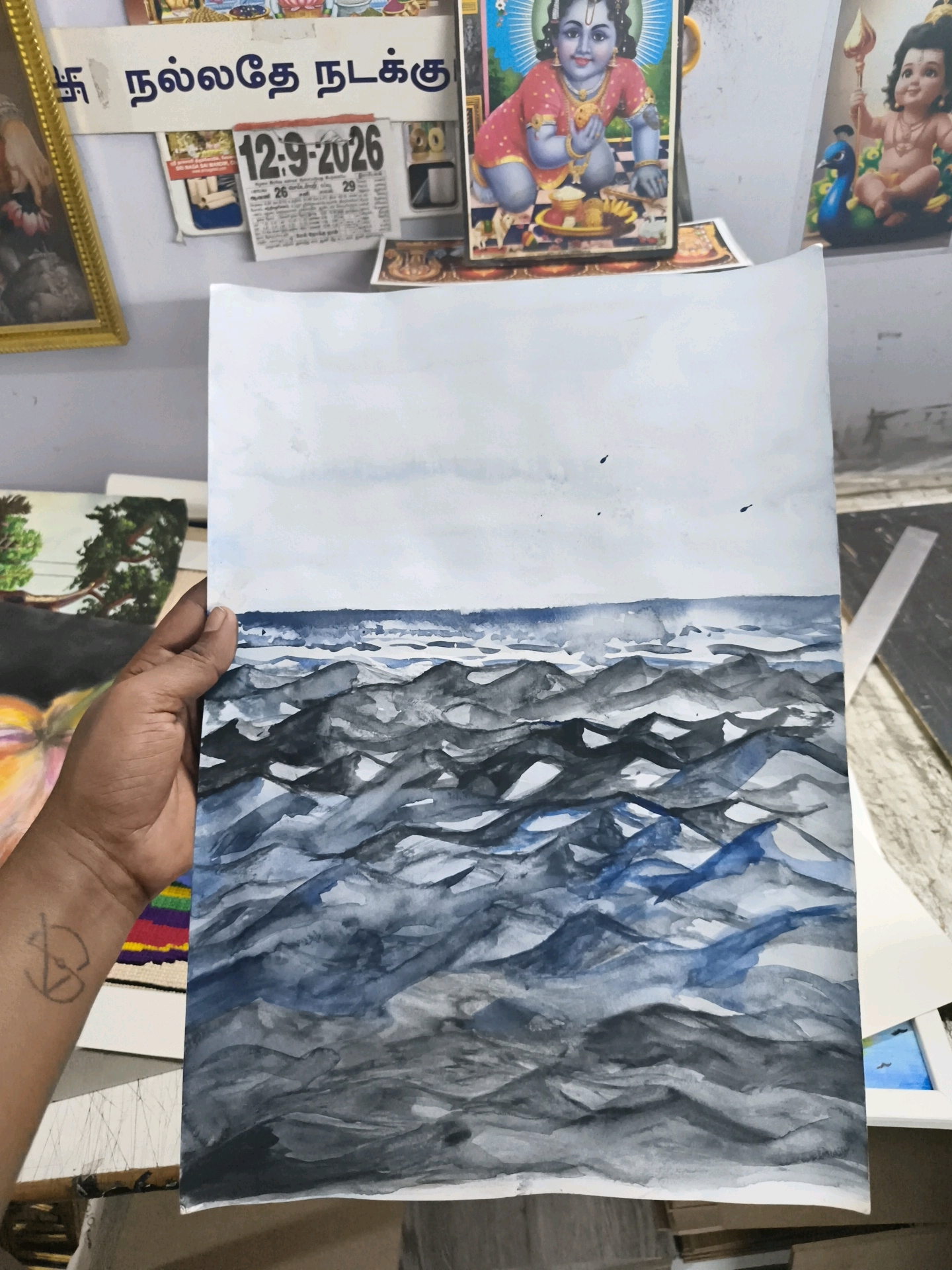 The image shows a close-up of a person's arm and hand holding a watercolor painting of a rough sea under a cloudy sky. The painting is predominantly in shades of blue, gray, and white, with dark, textured waves dominating the composition. A small tattoo is visible on the person's wrist. In the background, several posters and calendars are partially visible, including a calendar showing "12-9-2026" and a colorful image of a Hindu deity. The overall setting appears to be an indoor space, possibly a room or a workshop, with a plain wall as the backdrop. There is no discernible information to pinpoint the exact location in Coimbatore, India, nor any specific actions or interactions taking place beyond the act of holding the painting.