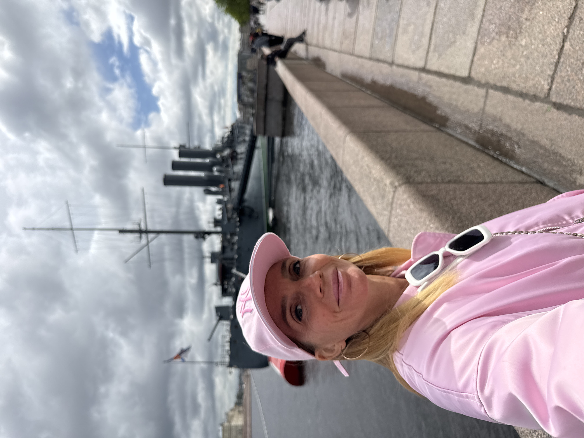 A woman wearing a pink hat and jacket is taking a selfie in front of the Cruiser Aurora in Saint Petersburg, Russia. The ship is docked on the Neva River and is visible in the background. The sky is cloudy. People are sitting on the embankment in the background.