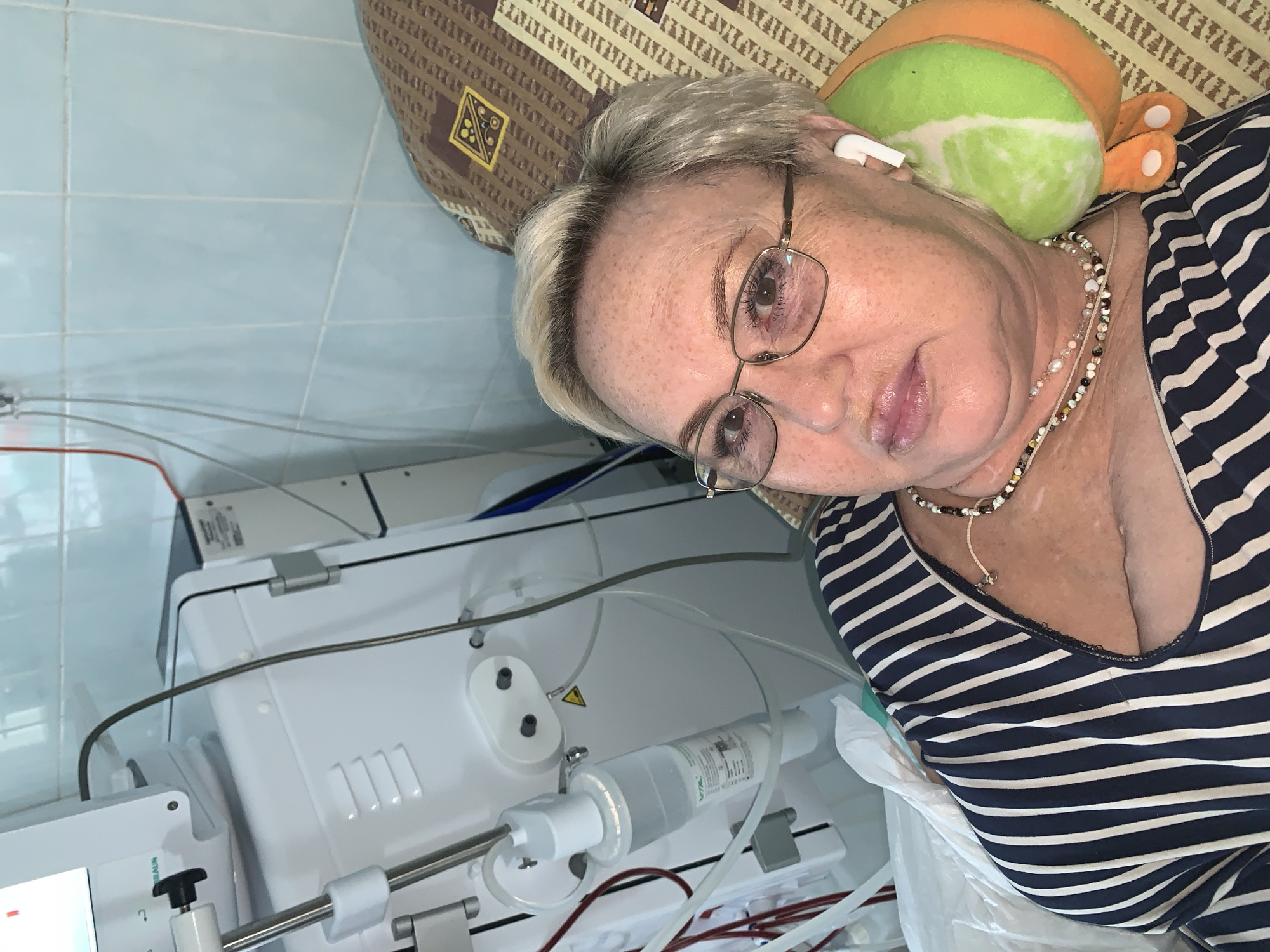 A middle-aged woman with short blonde hair and glasses is lying on her back, with a striped shirt, on what appears to be a medical bed or recliner. She has an orange and green plush toy under her head and is wearing earbuds. Next to her is a medical device, likely a dialysis machine, with visible tubing and containers. The background shows tiled flooring and a brown patterned cushion. The location and city of Chaikovsky, Russia are not determinable from the visual information provided in the image.