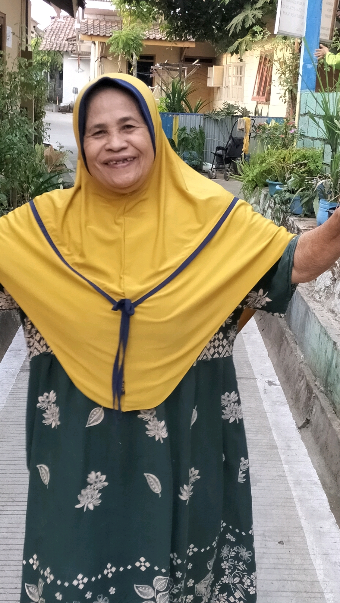 An Indonesian woman, appearing to be in her late 50s or early 60s, is standing outside, smiling broadly and looking directly at the camera. She is wearing a bright yellow hijab with a dark blue trim and a dark green dress adorned with a white floral pattern. Her arms are extended outwards, suggesting a welcoming or joyful gesture.
The background of the image depicts a residential area in Subah, Indonesia. There are houses with tiled roofs, some with visible windows and doors. Lush green plants and trees are scattered throughout the scene, some in pots and some growing naturally. A stroller is visible in the middle ground, and a painted fence runs along the right side of the image. The ground in front of the woman is paved with concrete, marked with white lines, indicating a walkway or street. The overall environment suggests a warm, sunny day.