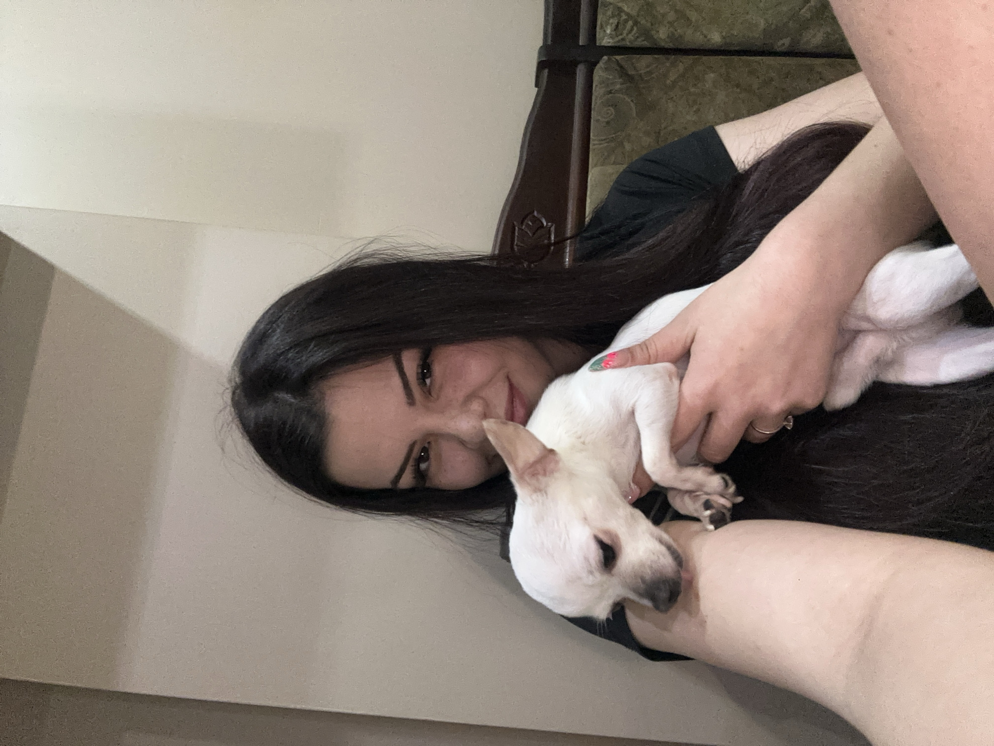 A woman with dark hair and light skin is holding a small, white dog. The woman is smiling and looking at the camera. The dog is nestled in her arms. The background is a light-colored wall and part of a dark-colored, possibly wooden, headboard. The location cannot be determined from the image alone. The city of Sliven, Bulgaria, cannot be confirmed.