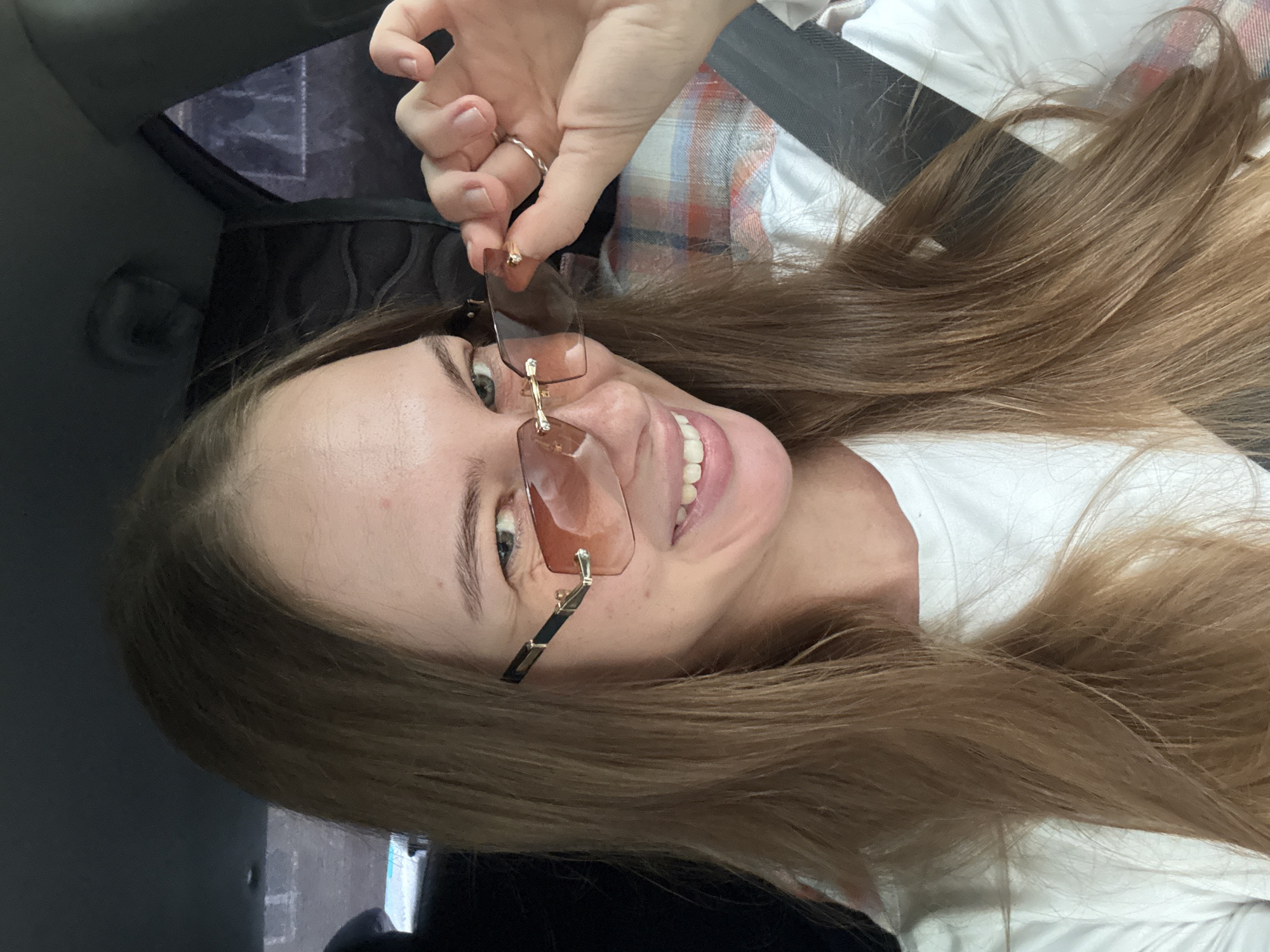 This media file is a close-up, vertically oriented photograph of a young woman with long, light brown hair. She is wearing rimless, light brown tinted sunglasses and a silver ring on her right index finger. She is smiling with her mouth open, revealing her teeth. Her skin appears fair. The background is blurred, but suggests an indoor setting, possibly a car, with dark upholstery and a hint of a plaid pattern on a seat or clothing. The lighting is bright, suggesting it is daytime, and there are no obvious clues about the weather. The overall emotion conveyed is one of happiness or amusement. There is no visible text in the image.