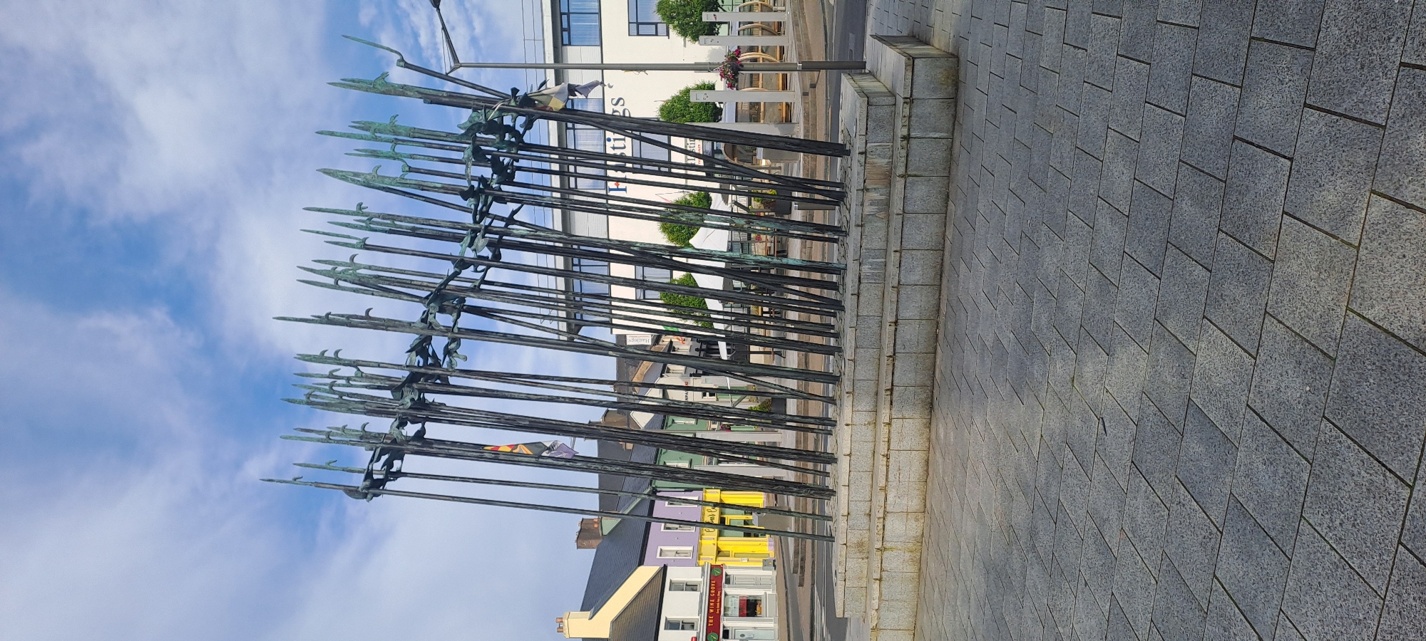 The image shows a public space in Castlebar, Ireland, featuring a sculpture composed of numerous tall, thin metallic poles with pointed tips, arranged in a semi-circular fashion. The sculpture is erected on a tiered stone base, which in turn is situated on a paved area made of grey rectangular tiles. In the background, there are several buildings, some painted in bright colors like yellow and purple, and others in white with prominent signage. A street with parked cars is visible, and beyond that, more buildings and trees. The sky above is blue with scattered clouds. The overall scene depicts an urban environment with a prominent artistic installation.