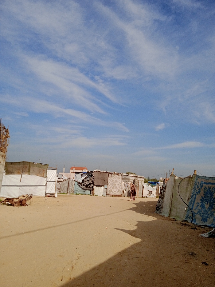 The image depicts an informal settlement in Khan Yunis, Palestinian Territory, under a bright blue sky with wispy white clouds. A wide, unpaved dirt path, light brown in color, occupies the foreground and recedes into the midground. Dark shadows from an unseen object to the right extend across the path. On both sides of the path are multiple makeshift structures, likely shelters, constructed from a variety of materials including corrugated metal sheets, wooden panels, white tarpaulins, and large, patterned fabric rugs or blankets serving as exterior walls.
Specifically, on the left, a structure is partially covered with white sheeting. Further along the left and center, a row of shacks features walls made from dark patterned rugs and lighter-colored patterned textiles. One individual, dressed in dark clothing, walks away from the viewer along the central path, roughly in the middle distance between the structures. On the right, another structure is draped with a blue and white patterned rug. In the distant background, beyond the informal settlement, more permanent buildings with light-colored walls and terracotta-colored roofs are visible against the horizon.